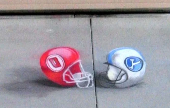 Two football helmets, one red and one white, with face masks, placed on a concrete surface facing each other.
