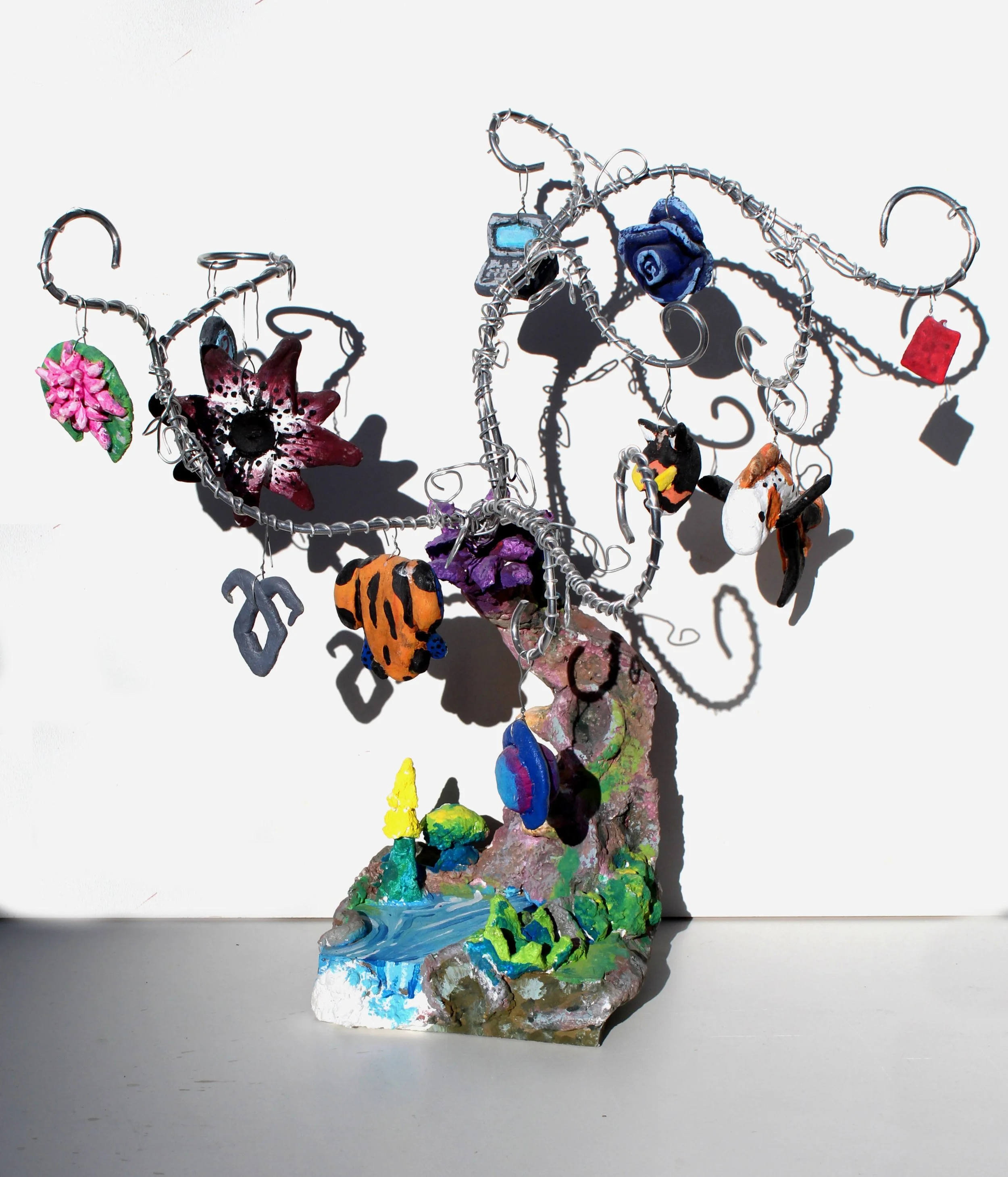 Colorful handmade sculpture of a whimsical tree with various charms and ornaments hanging from its branches, set against a plain white background.