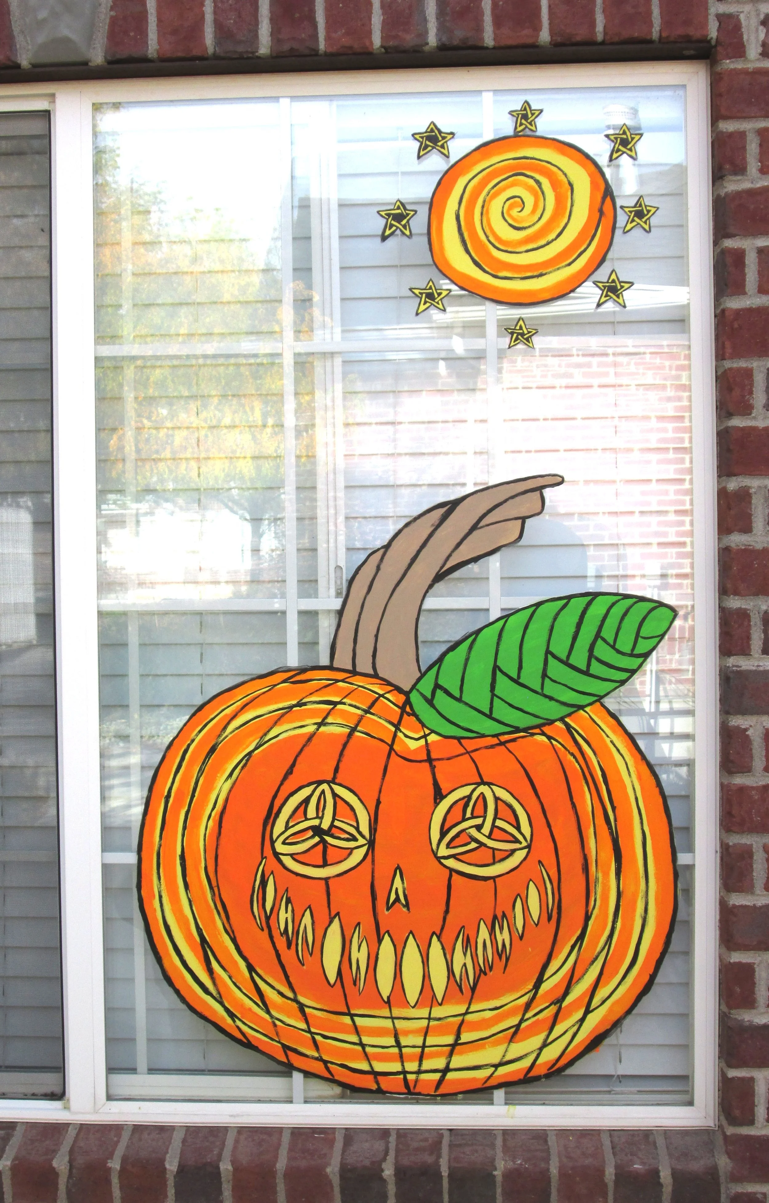 Window with Halloween-themed artwork of a large carved pumpkin with intricate designs, a spiral orange swirl at the top, and stars around it, painted in bright orange, yellow, and green colors.