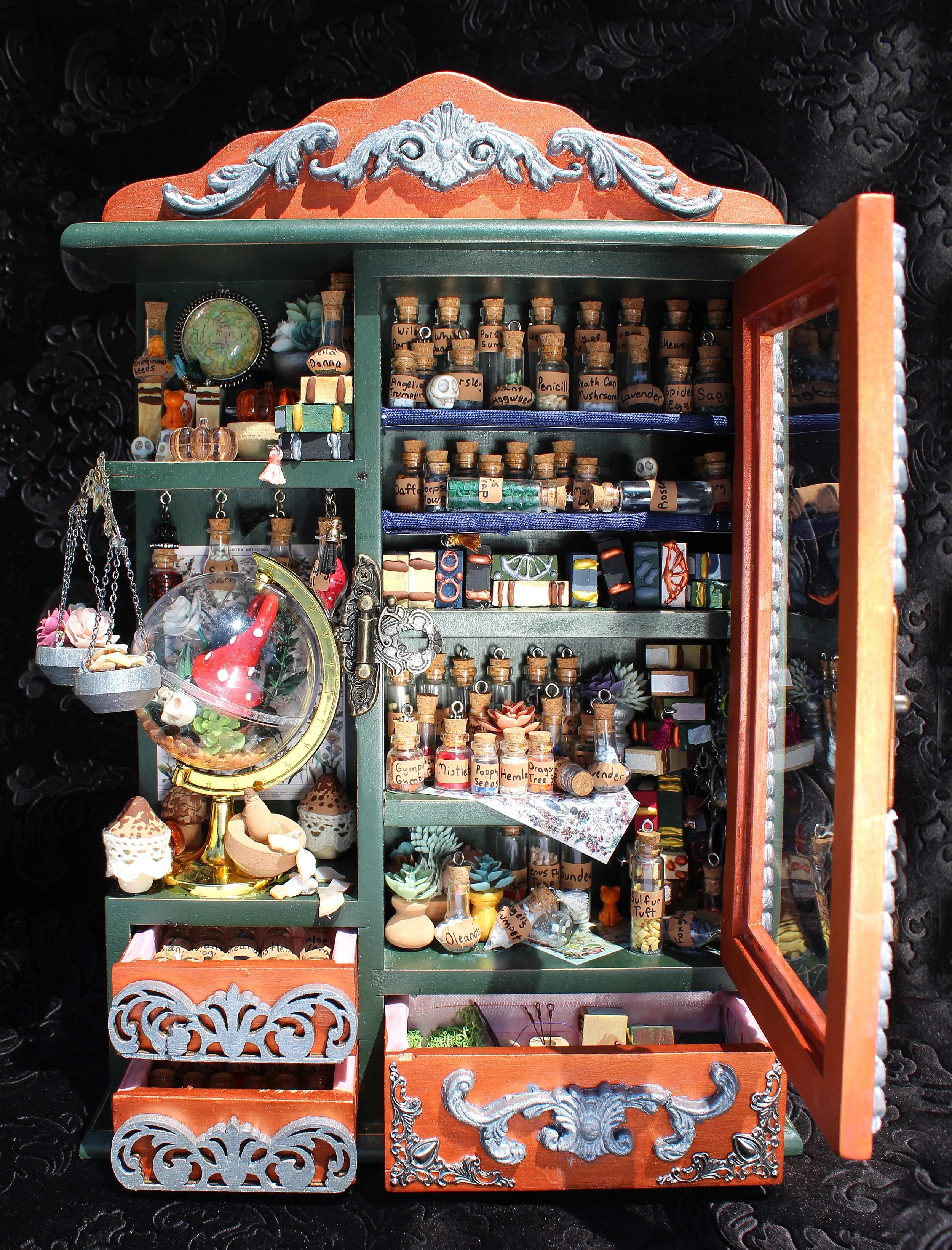 Miniature wooden cabinet with small glass bottles labeled with botanical names, decorative objects, and miniature accessories, displayed on a black textured background.