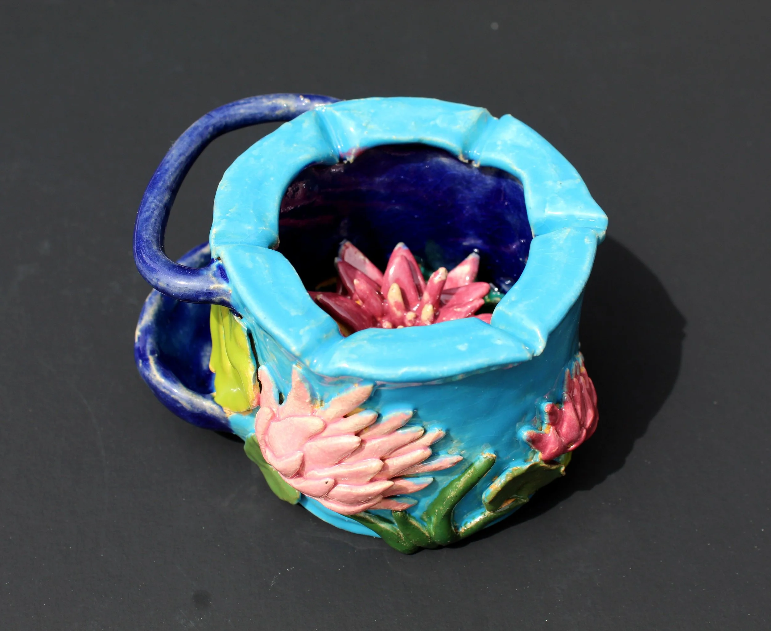 Colorful ceramic mug with a blue rim, featuring pink and green floral patterns, and containing a pink flower arrangement, set against a black background.
