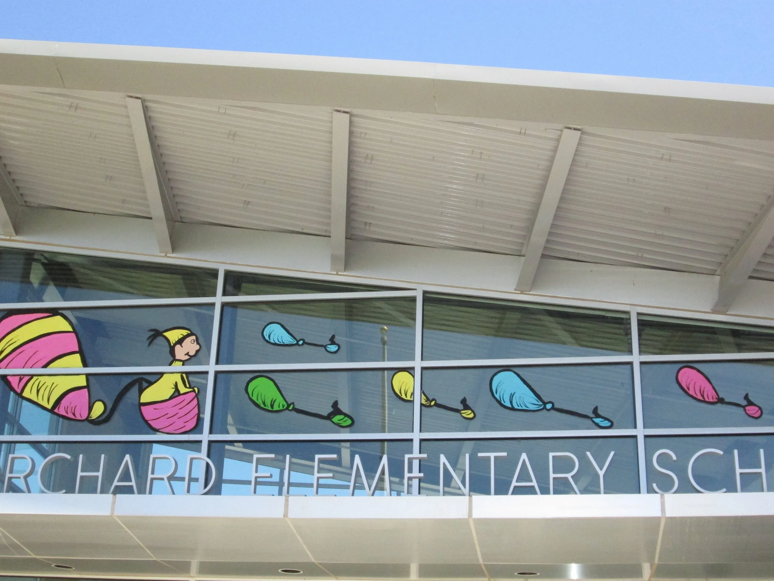 Exterior view of a building with large windows decorated with colorful yarn art featuring knitting needles and balls of yarn, and a partial sign reading 'SCHOOL OF CHARD ELEMENTARY SCHOOL'.