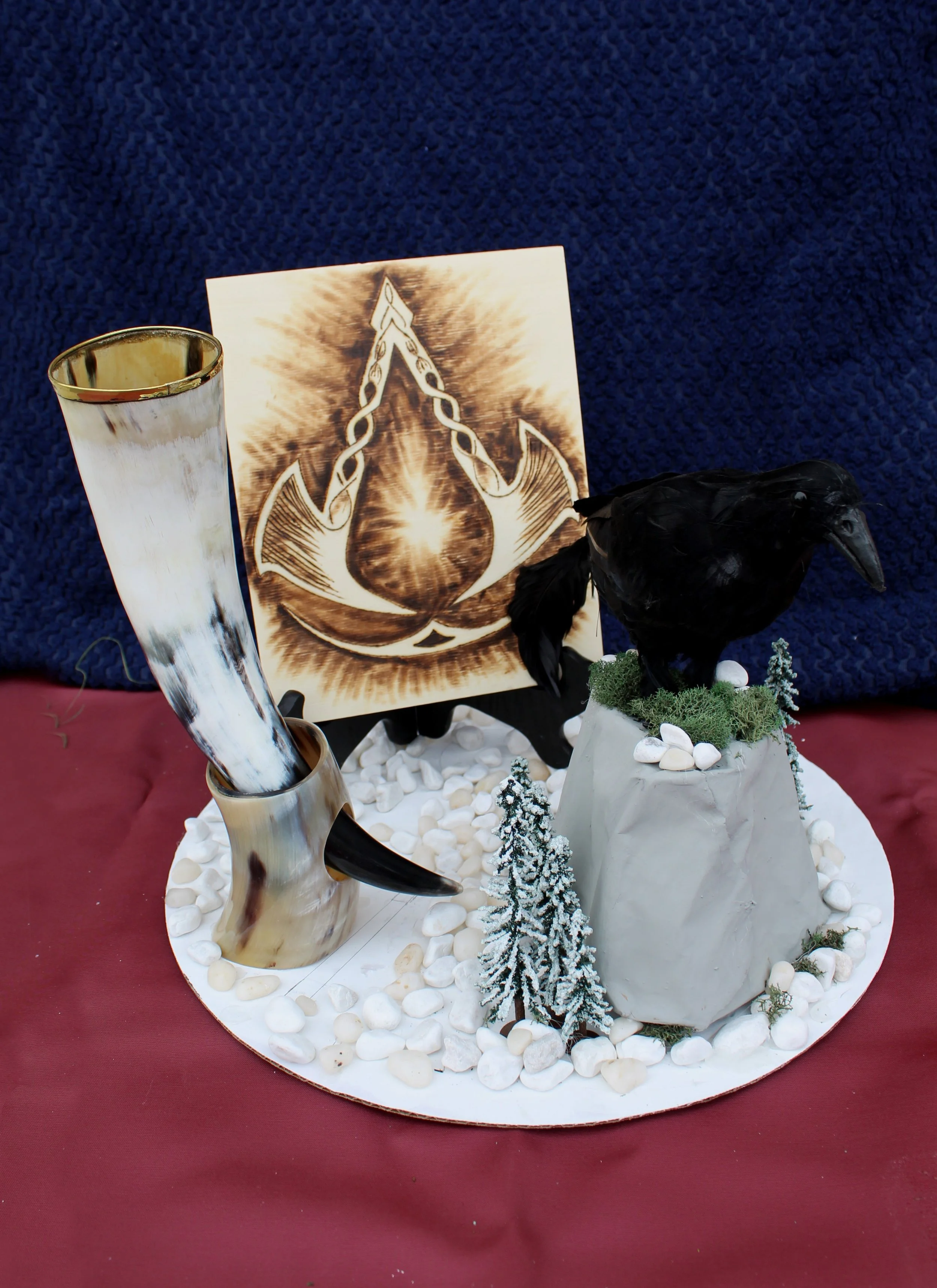 A decorative display with a horned black bird sculpture, a tall glass with a white and brown swirl pattern, a picture with a Viking ship icon, and small white rocks and miniature trees arranged on a circular white base against a blue textured backgro