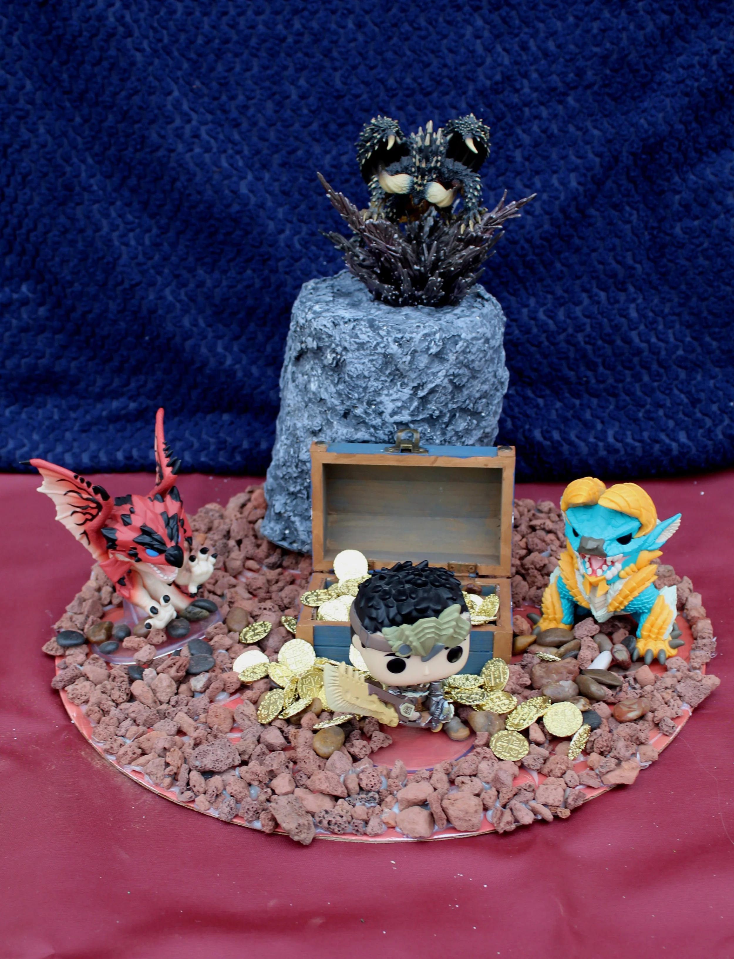 Display of dragon figurines with a treasure chest filled with gold coins, surrounded by rocks and decorative elements, set on a round red table with a dark blue textured background.