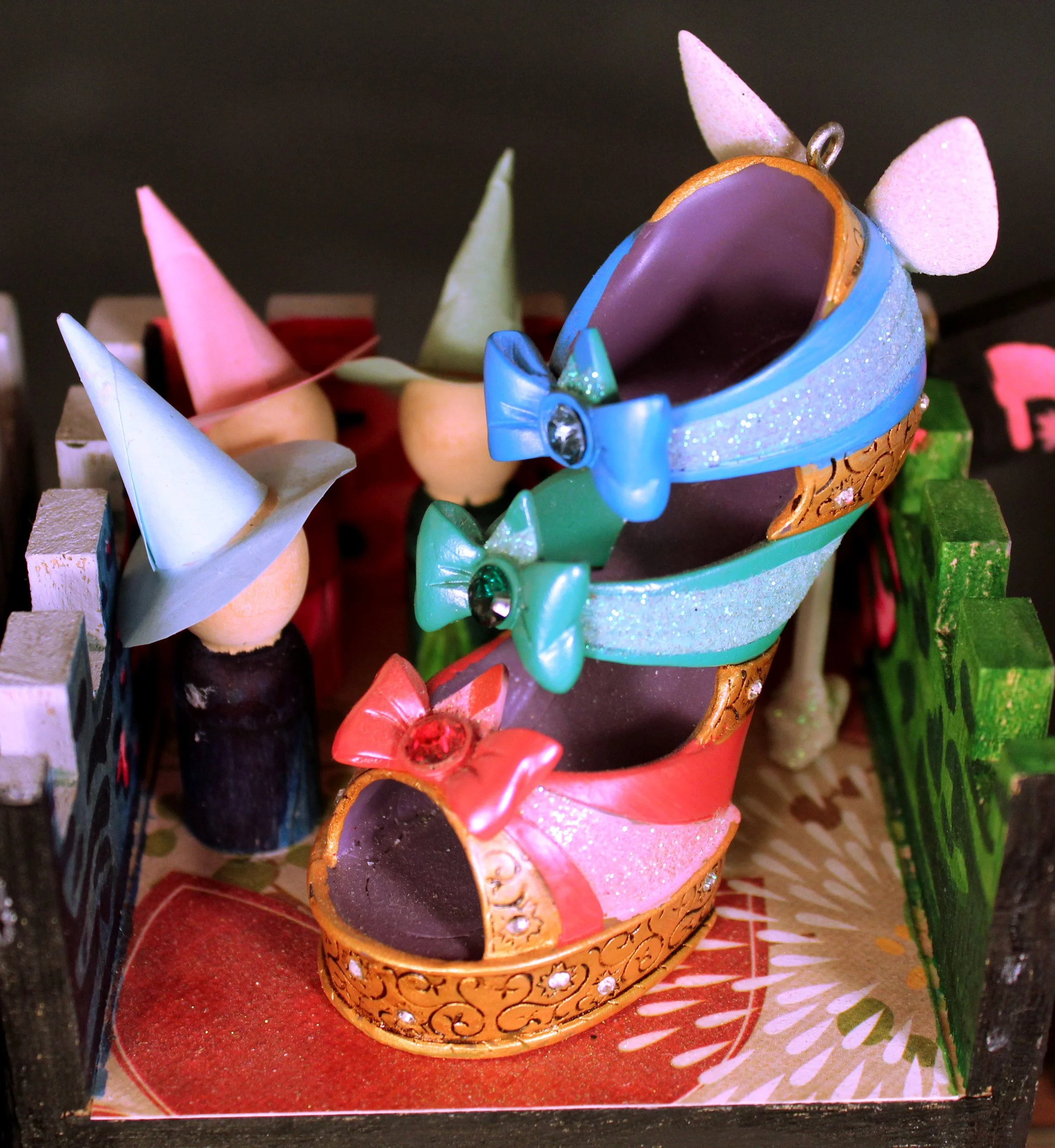 A festive, colorful cone-shaped cake decorated to look like a unicorn, with pink, purple, and blue icing, a glittery surface, and pink and white unicorn ears and horn. The cake is surrounded by other decorated cakes and desserts.