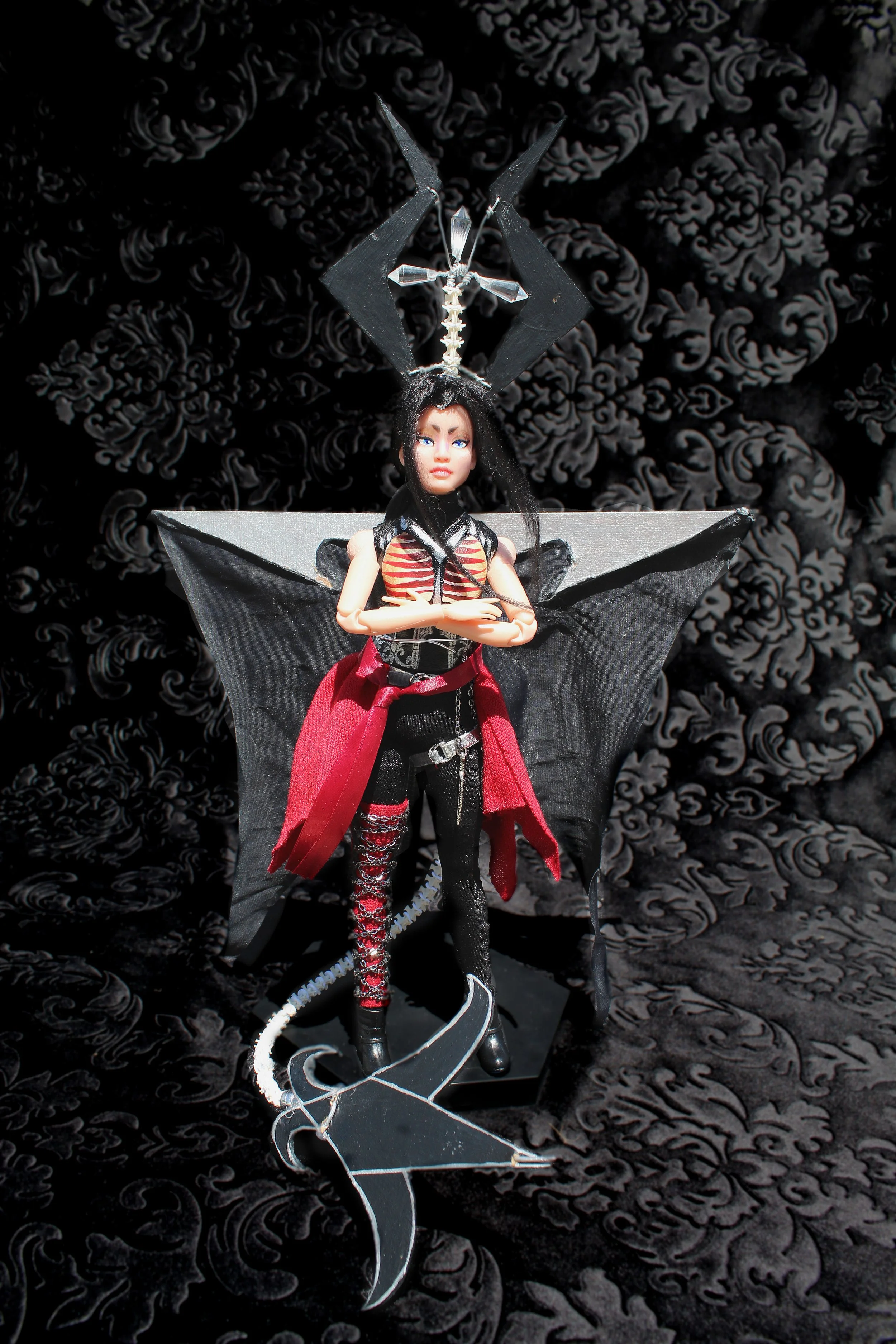 A doll dressed in punk-style clothing with black and red colors, standing in front of black floral-patterned background.