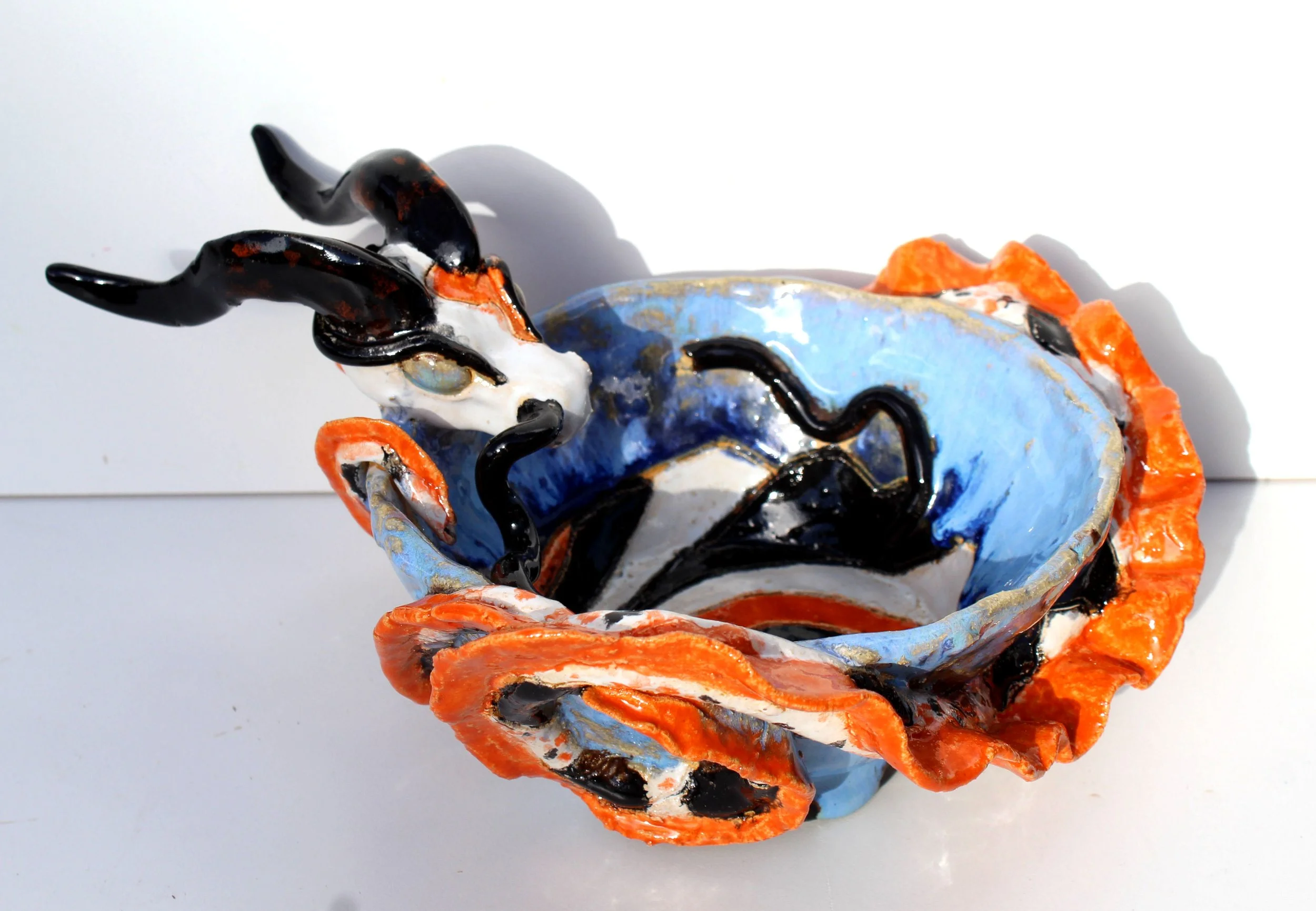 Close-up of a ceramic sculpture resembling a creature with a blue body, black lines, orange accents, and black horns, set against a white background.