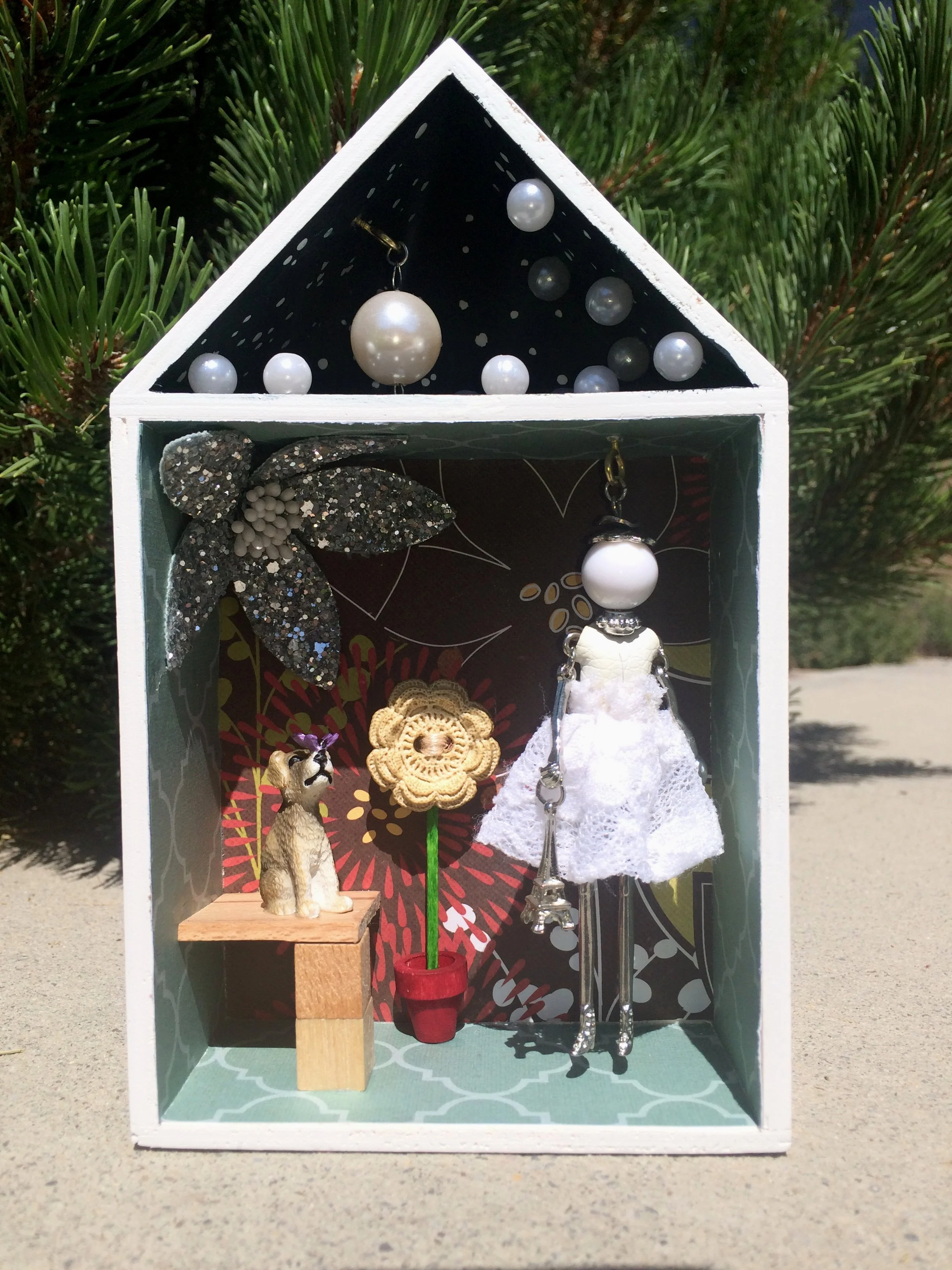 Decorative miniature house display with various hanging and standing jewelry and accessories, including pearl jewelry, a black glittering leaf, small dog figurine, and a flower-shaped earring on a wooden block, set outdoors against a greenery backgro