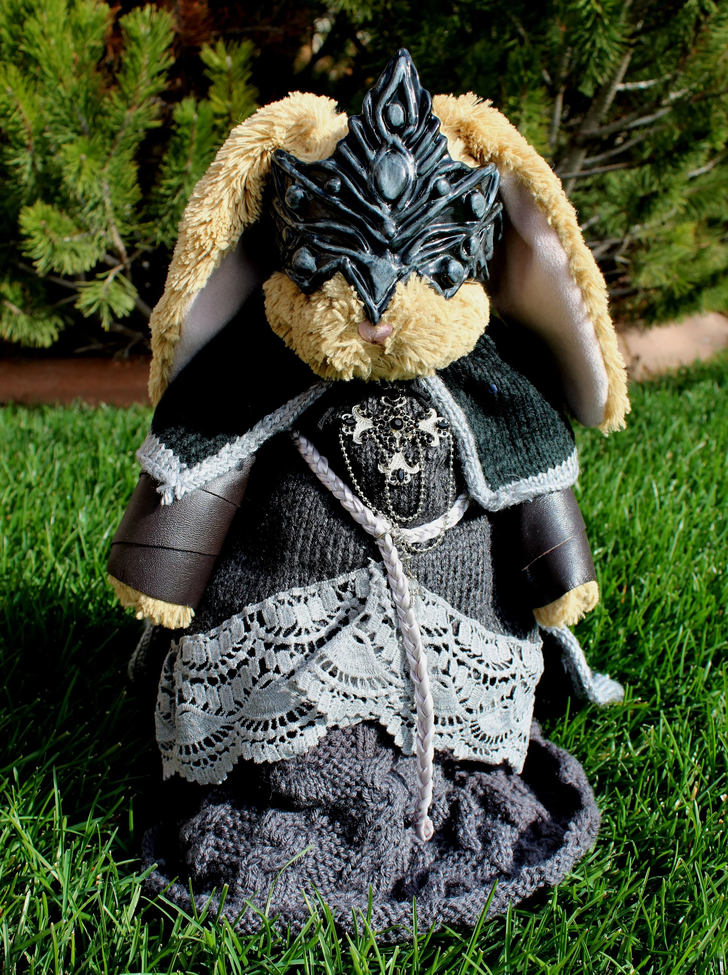 A doll with a rabbit costume and an intricate mask on its face, standing on grass with green shrubs in the background.