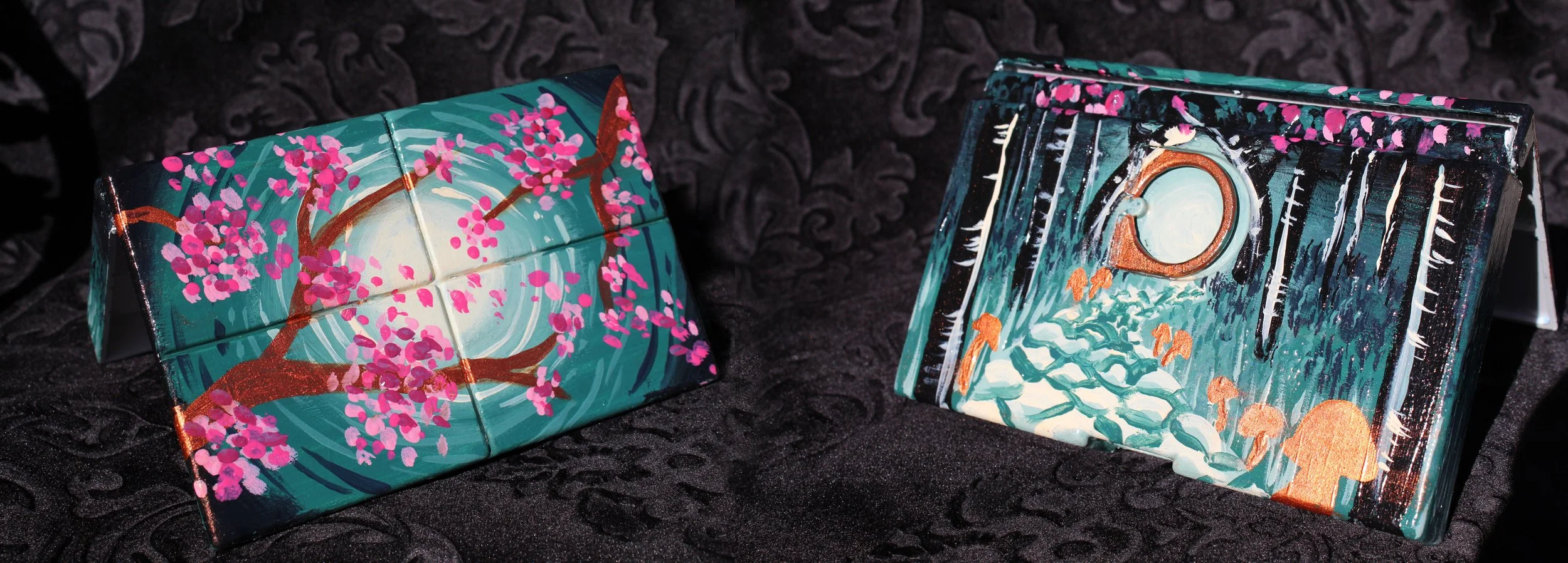 Two hand-painted, decorative boxes with nature-themed designs on black textured fabric background.