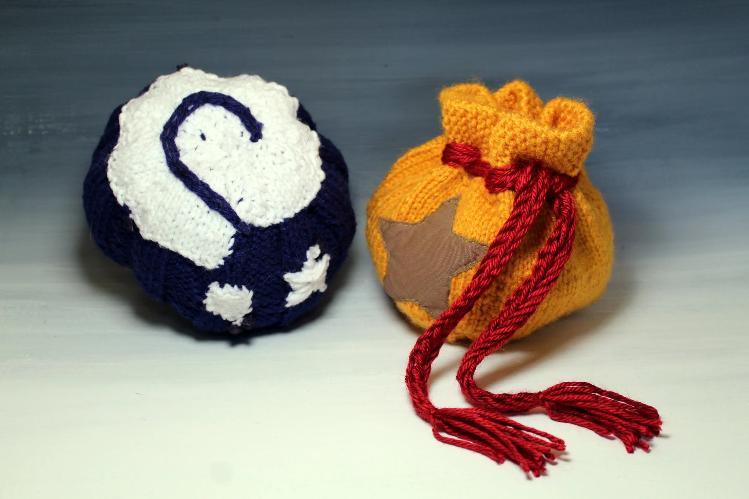 Two small knitted/patched objects resembling sock-like shapes, one with a white and navy blue design resembling a penguin and the other in a yellow-orange color with a star patch and red yarn ties, placed on a light blue surface.