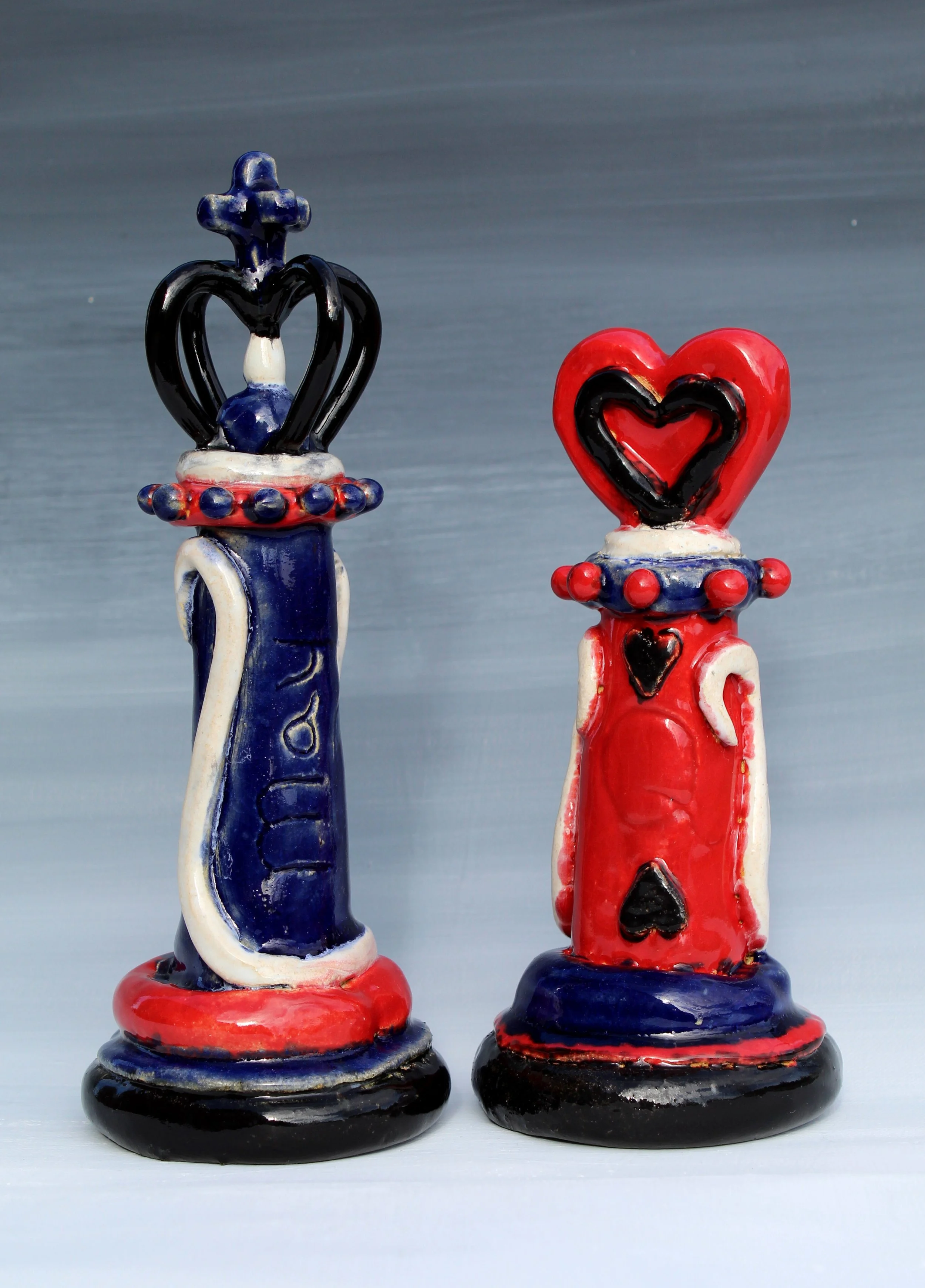 Colorful ceramic chess king and queen pieces, red and blue, decorated with heart and spade suits, placed on black and red bases against a gray background.
