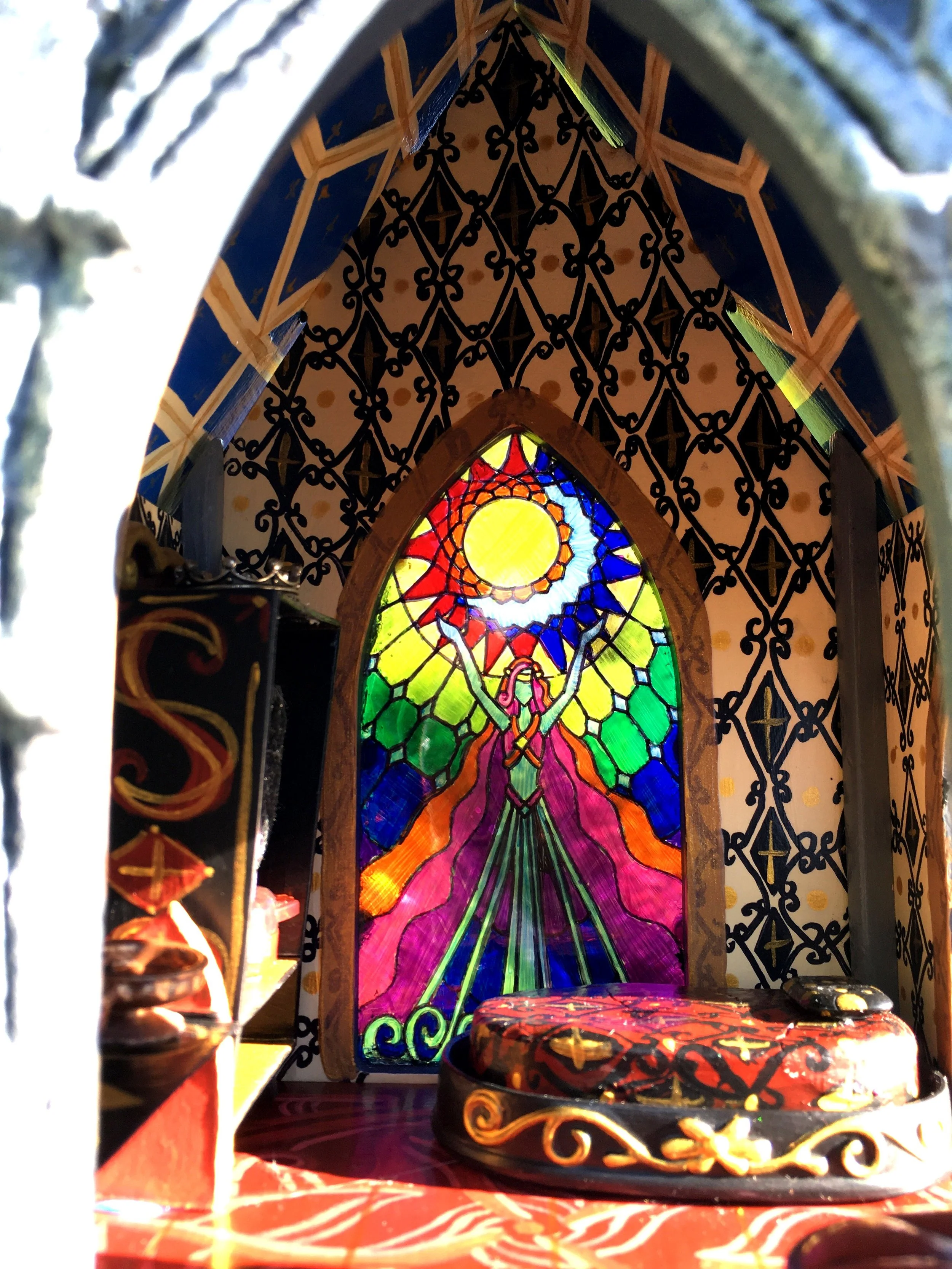 Inside of a decorative, colorful lantern featuring stained glass artwork of a woman in a pink dress against a multicolored background with a bright yellow sun at the top. The lantern has black metal framing with intricate patterns and is set on a sur