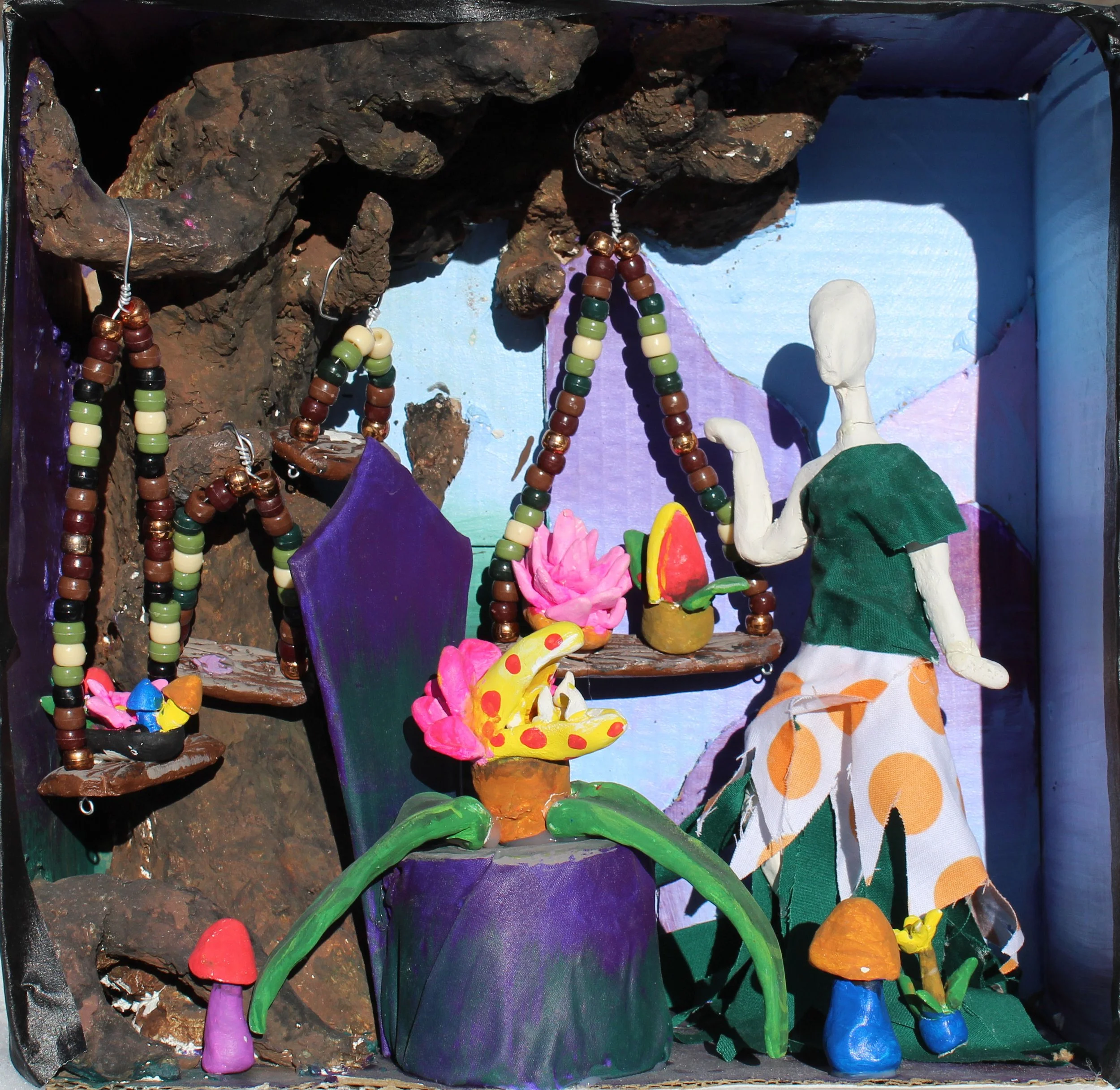 A diorama scene with colorful clay figures, including mushrooms, a creature resembling a caterpillar or snake, and a human figure with a green and orange polka dot dress, set against a painted background with rocks, flowers, and a blue sky.