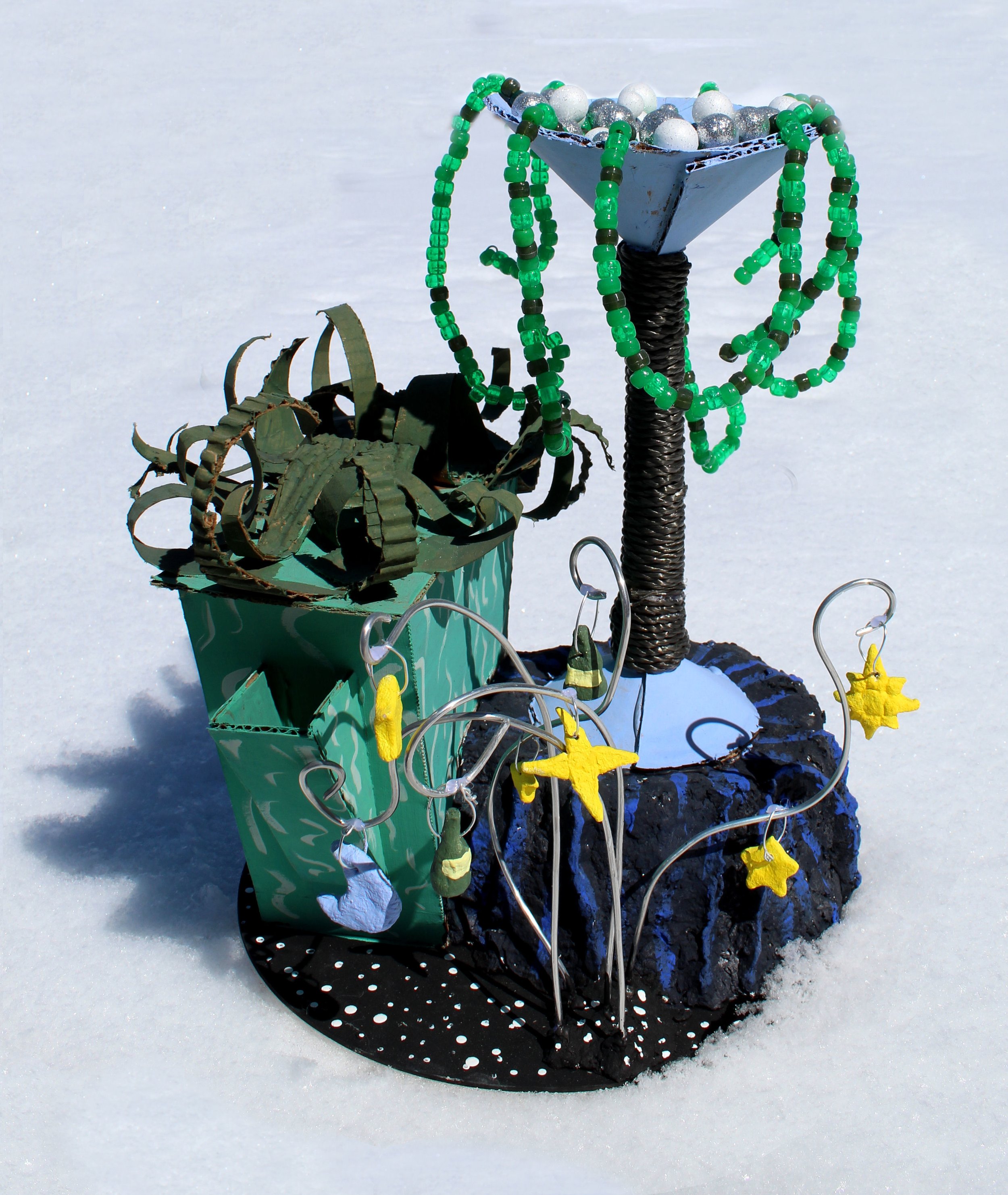 A festive holiday decoration with a black, polka-dotted, circular base and a small, green, rectangular container filled with metallic, silver, and white spherical ornaments. Several strands of green and black beads drape over the container and across