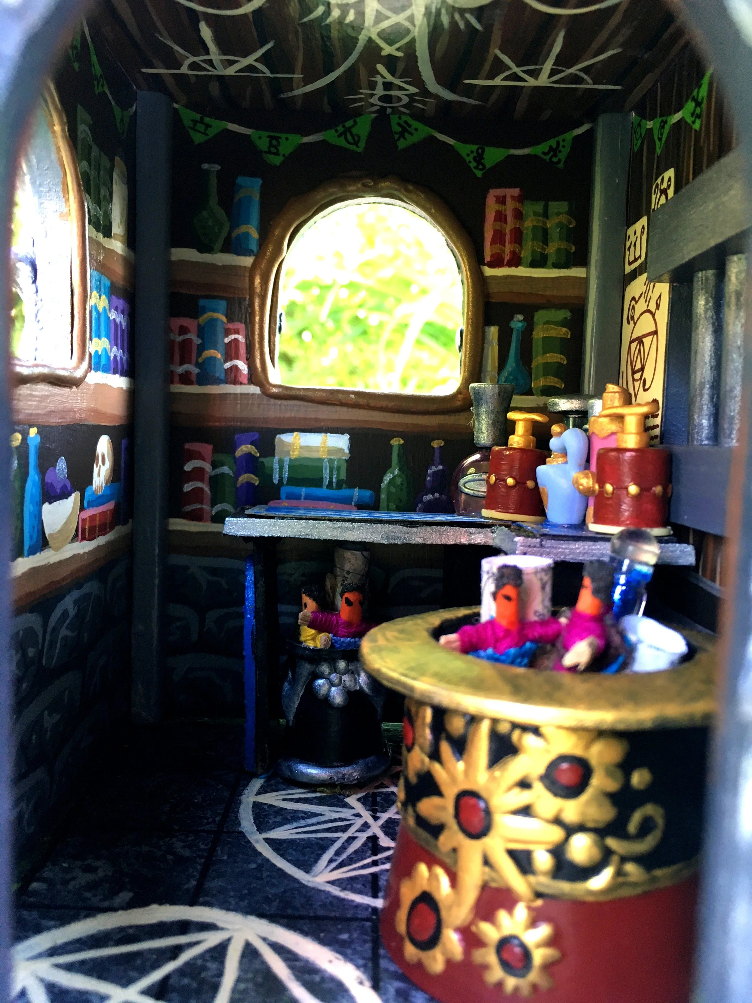 Miniature wooden room decorated with colorful painted books, bottles, and figurines, viewed through a small opening with a decorative floor pattern.