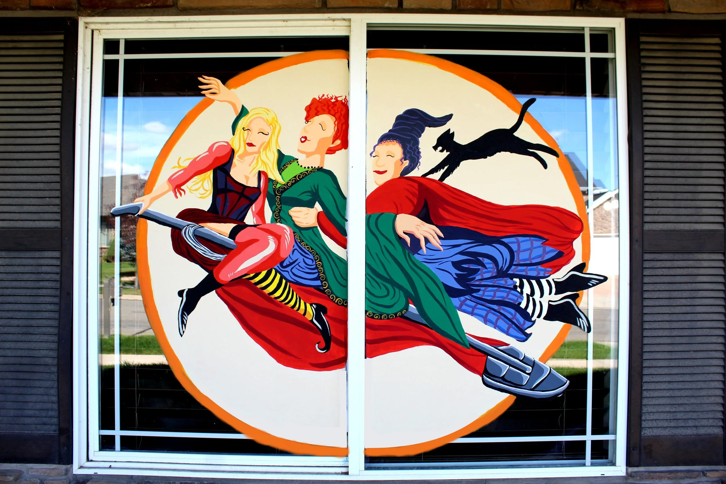 A colorful mural of three women riding a broomstick, with a black cat flying behind them, painted on a window with black shutters on either side.
