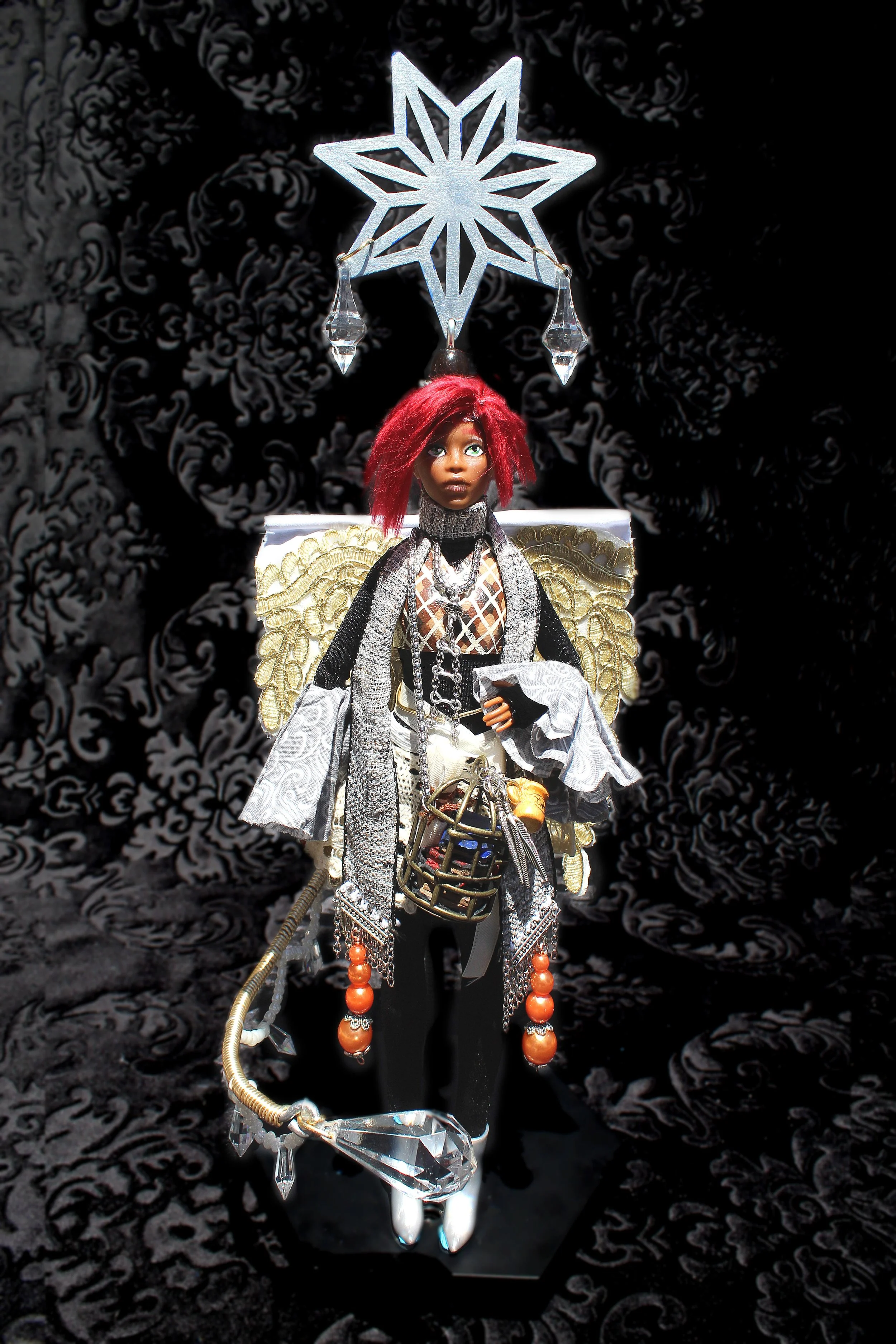 A doll with red hair dressed in elaborate fantasy or cosplay attire, holding a staff with a large crystal at the end, standing in front of a black and white patterned background and a decorative star-shaped hanging ornament above.