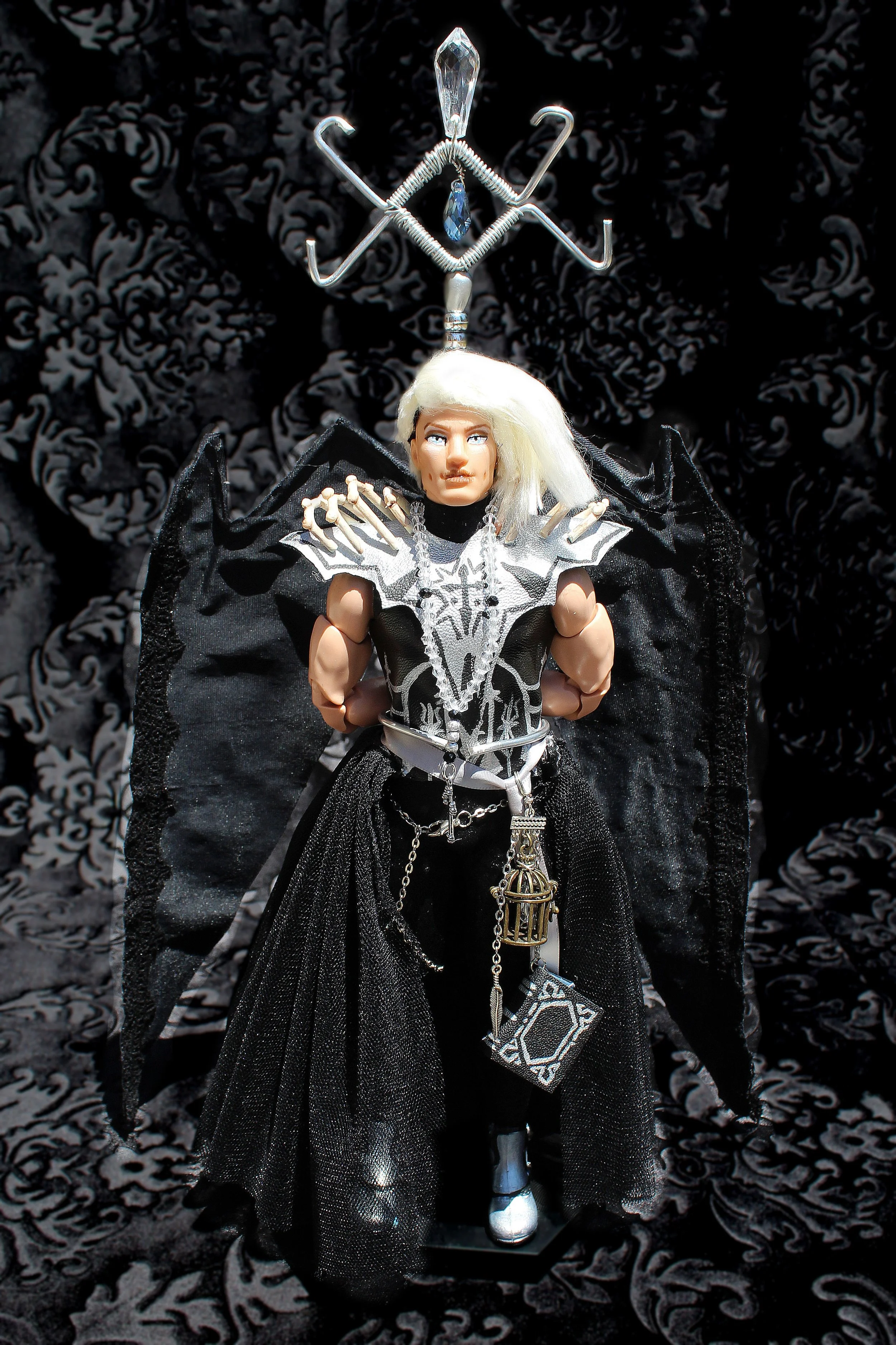 A doll dressed in black and silver gothic-style clothing, with white hair, standing against a black and white damask-patterned background, featuring an intricate headpiece with silver wires and crystal accents.