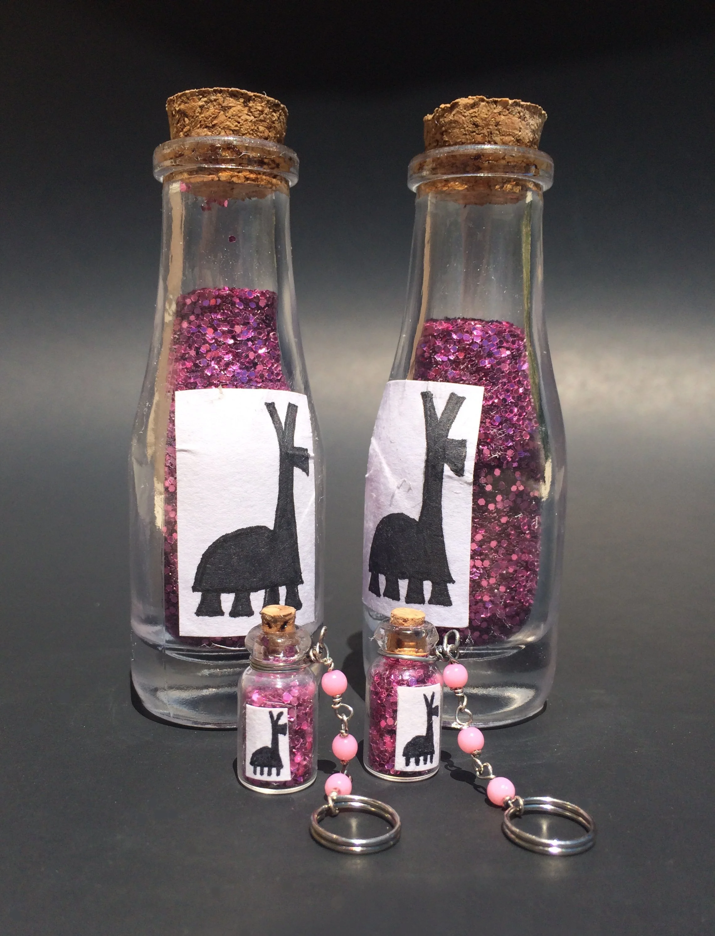 Two glass vials filled with pink glitter, each sealed with a cork, have labels with a silhouette of a black pig and the letter 'Y'. Two small glass jars with pink beads are attached to the bottom of each vial with small chains.