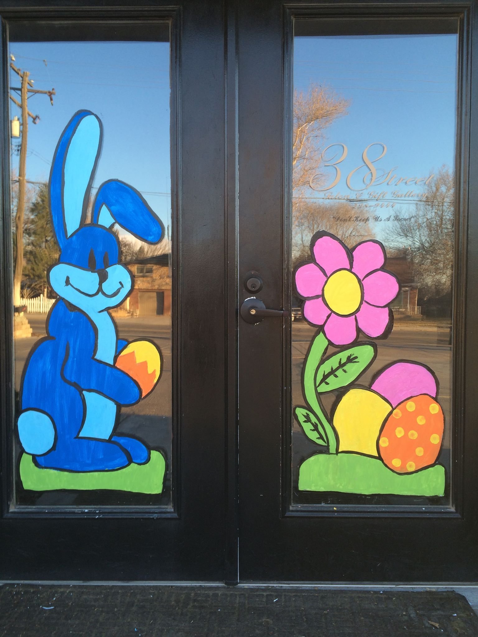 Decorative glass door with colorful illustrations including a blue cartoon rabbit holding a yellow and orange egg on the left and a pink flower with green leaves and orange egg on the right. The door has a black frame with a handle and lock. Text on 