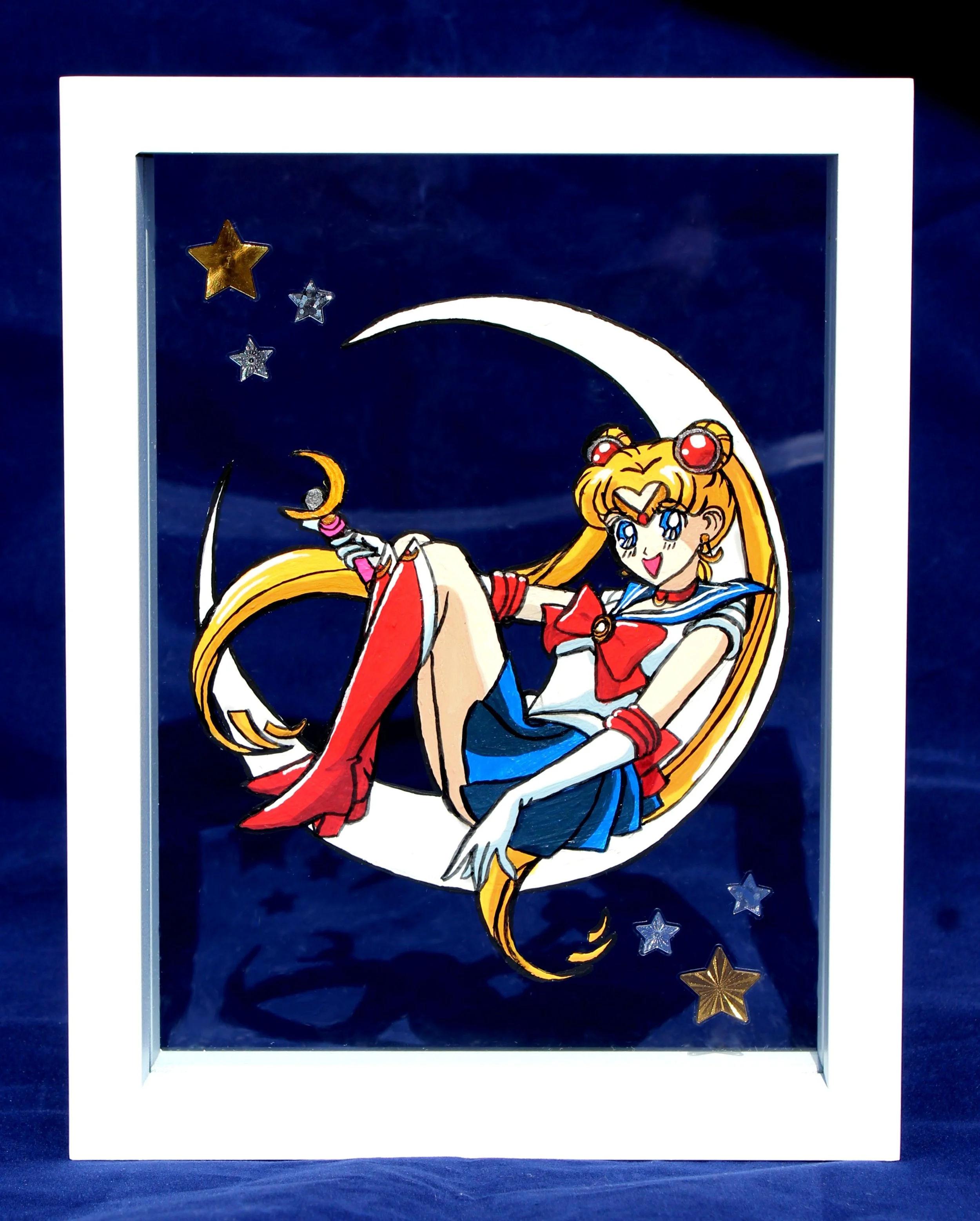 A framed illustration of Sailor Moon with her blonde hair and blue and white sailor outfit, holding a crescent moon staff, surrounded by stars and a dark blue background.