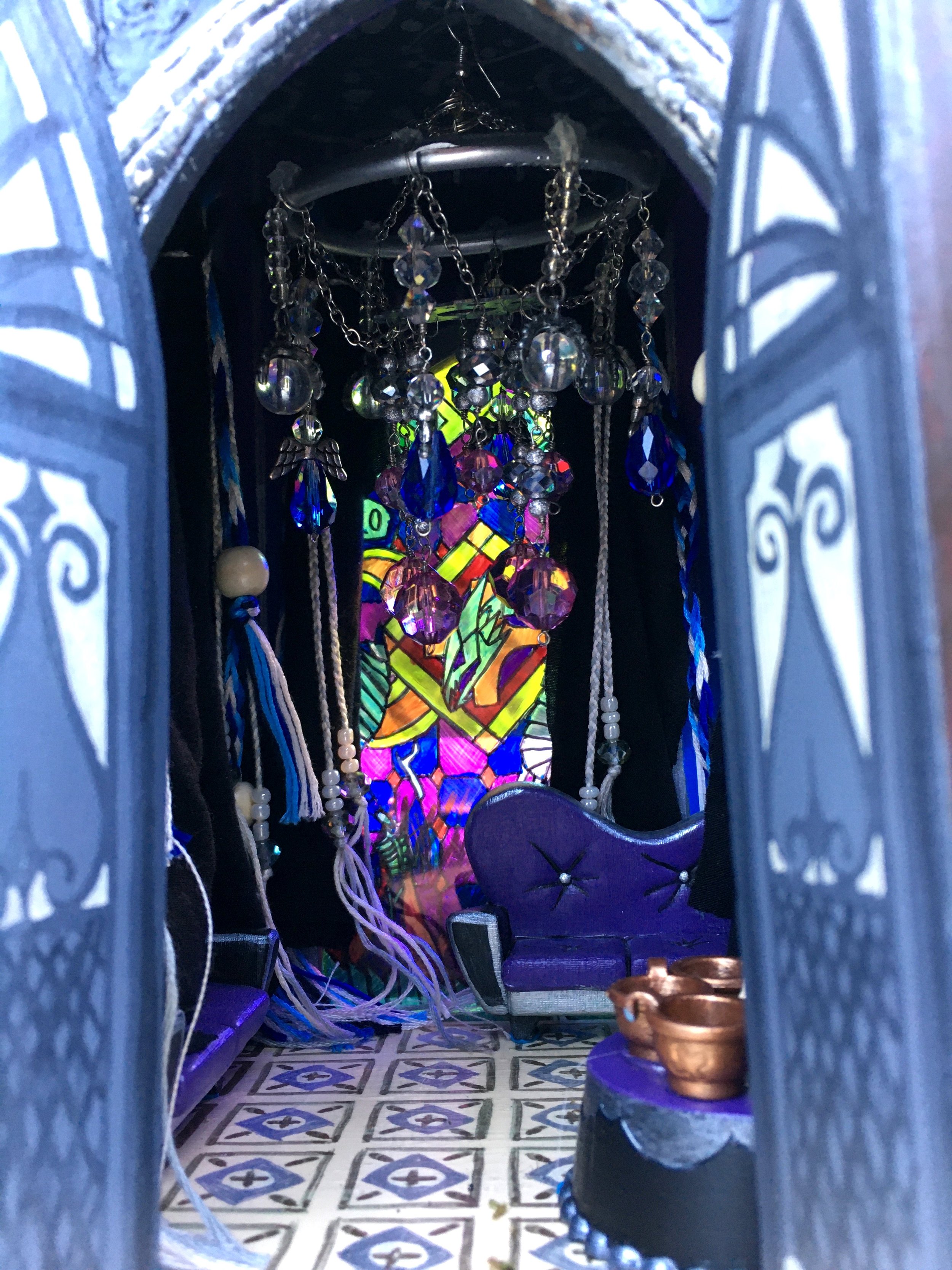 A view inside a small, ornate church or religious shrine, featuring hanging crystal decorations, a colorful stained glass mural behind, and a patterned tile floor.