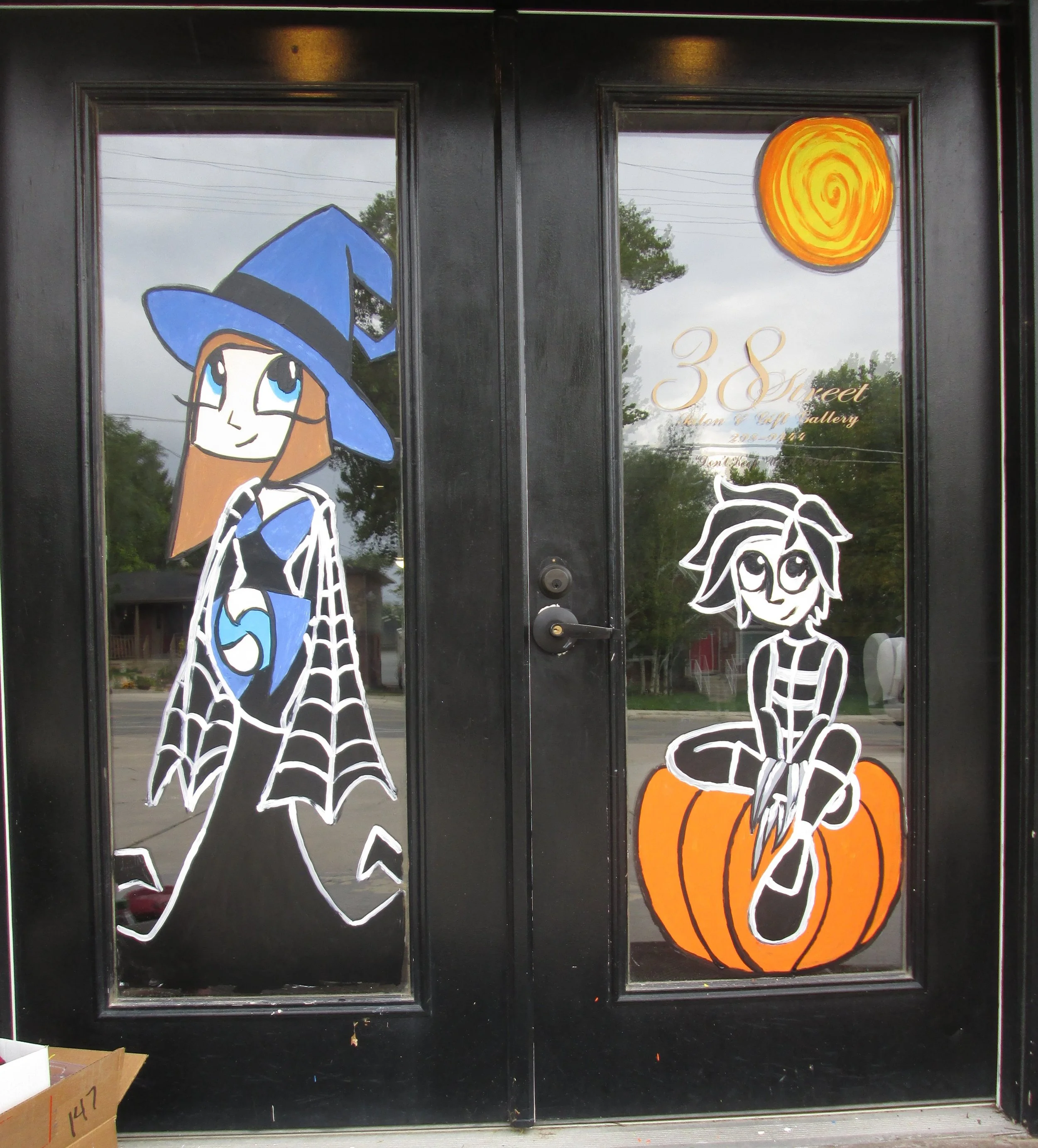 Window decals of a girl with a blue witch hat and black and white dress on the left, a bright yellow sun on the upper right, and a black and white cat with pumpkin on the bottom right.