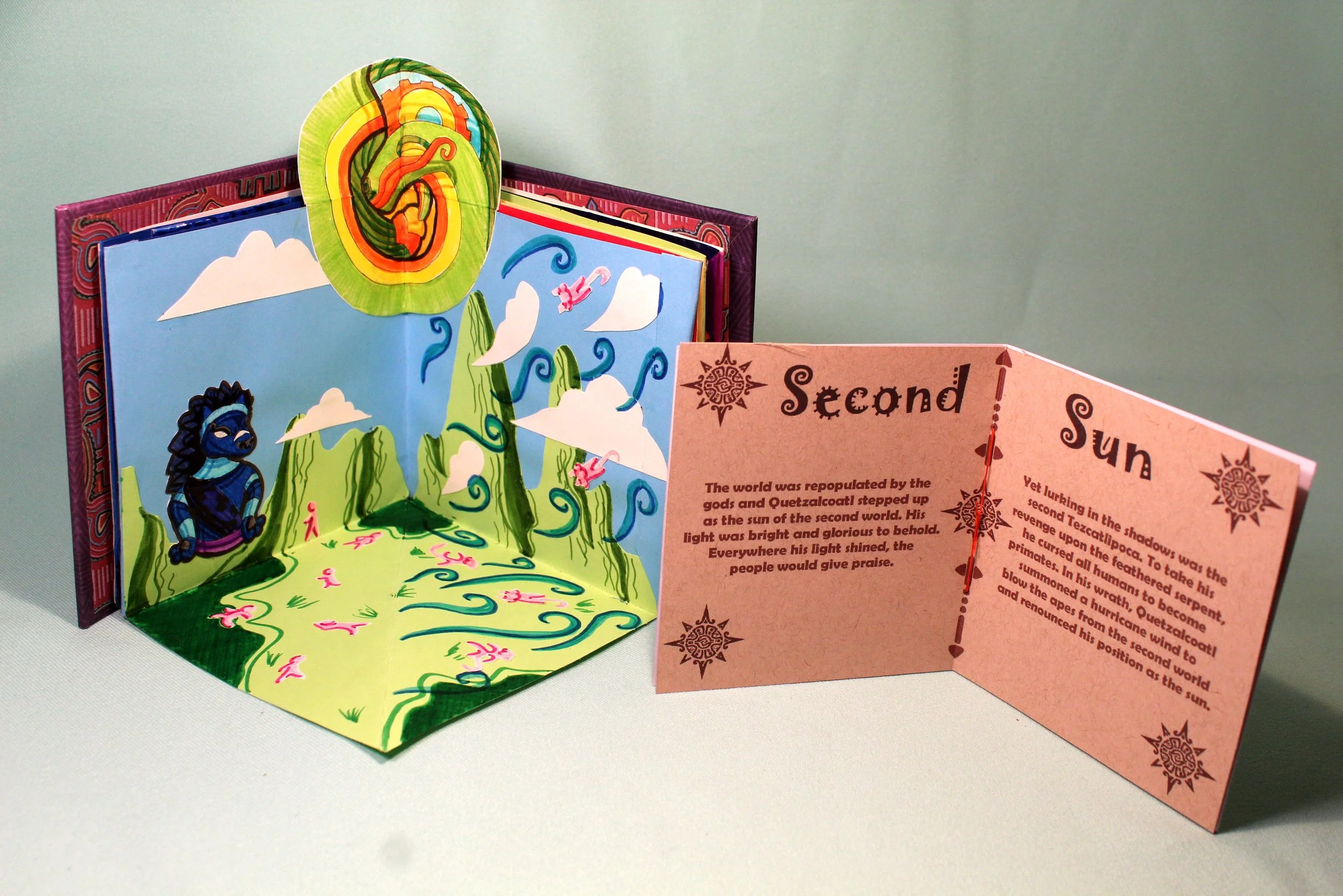 Open pop-up book with colorful illustrations and a pink folded page with text, depicting a fantasy scene with a mountain, clouds, and a blue lion character.