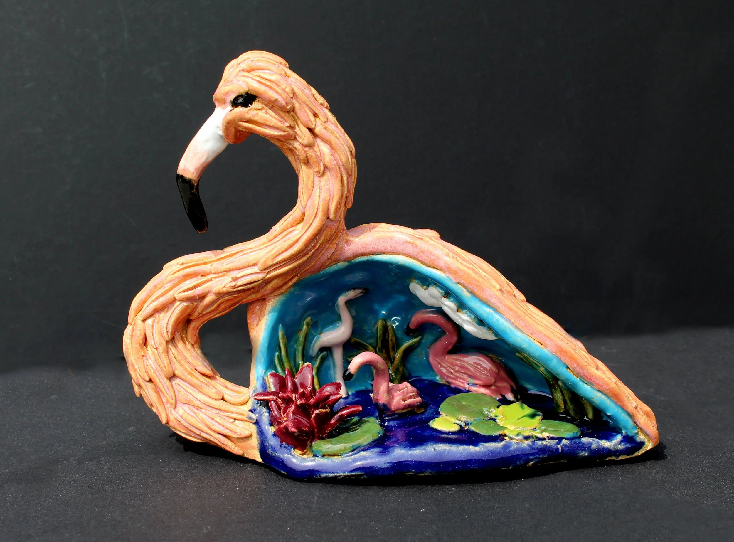 Colorful ceramic sculpture of a flamingo with a hollow interior filled with small decorative elements, set against a black background.