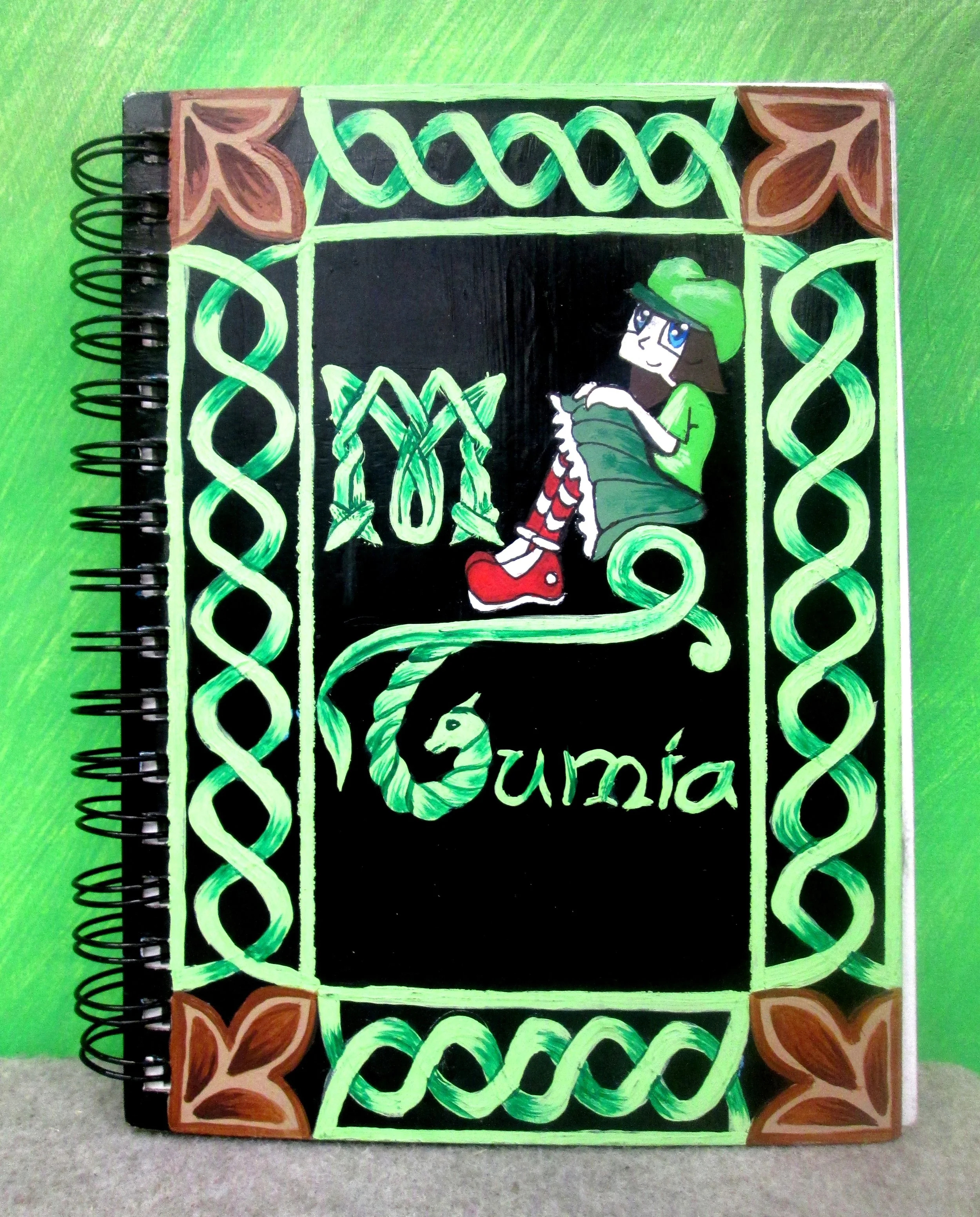Spiral-bound notebook with a hand-drawn colorful cover. The cover features a green border with brown corner decorations, green vine-like patterns, and the words "Mi Quimía" written in green. There is a cartoon girl with long brown hair, blue eyes, we