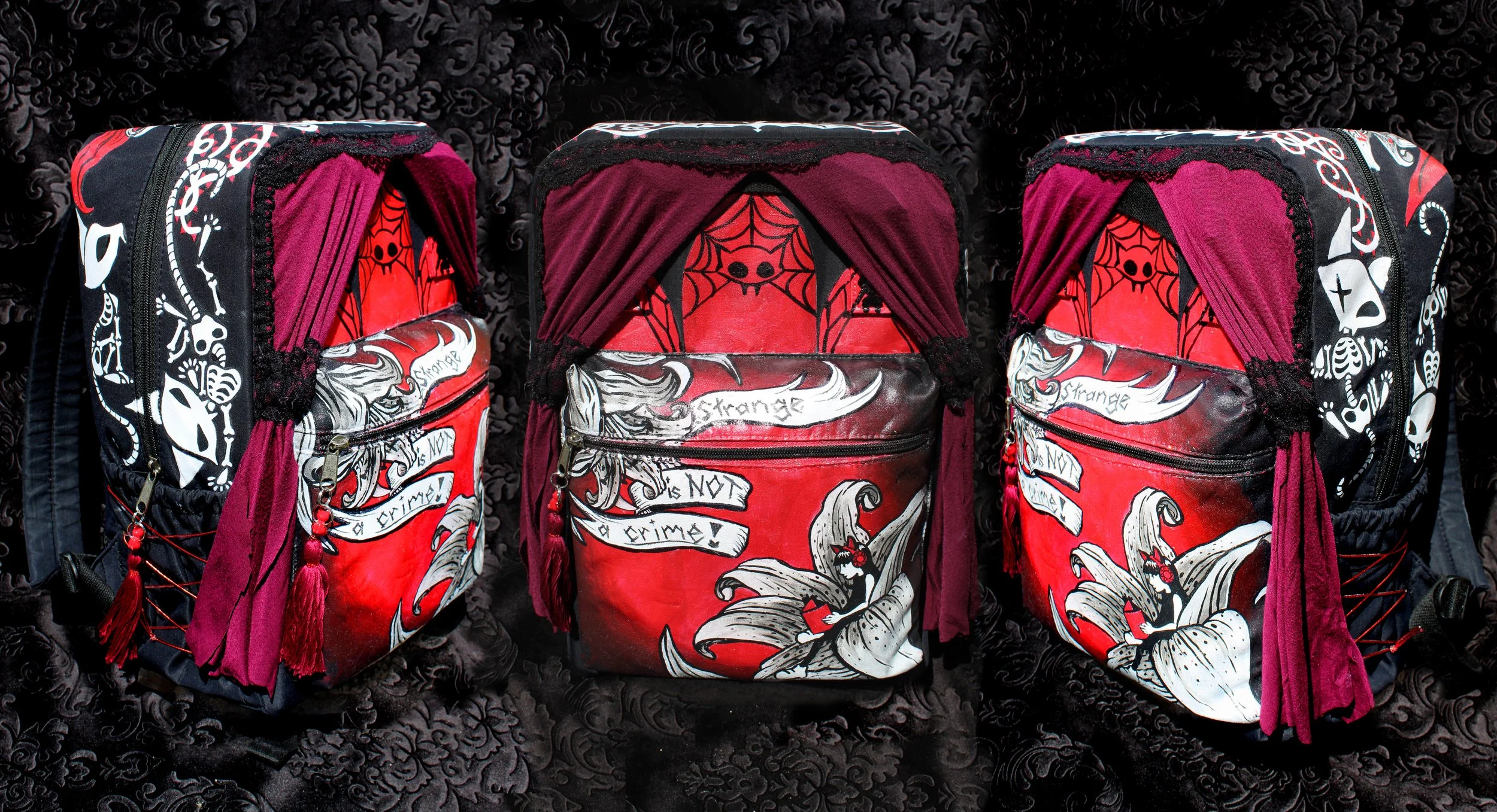 Three backpacks with red, black, and white skull, spider web, and tattoo-style designs, featuring dark backgrounds and maroon accents, displayed on a black textured surface.