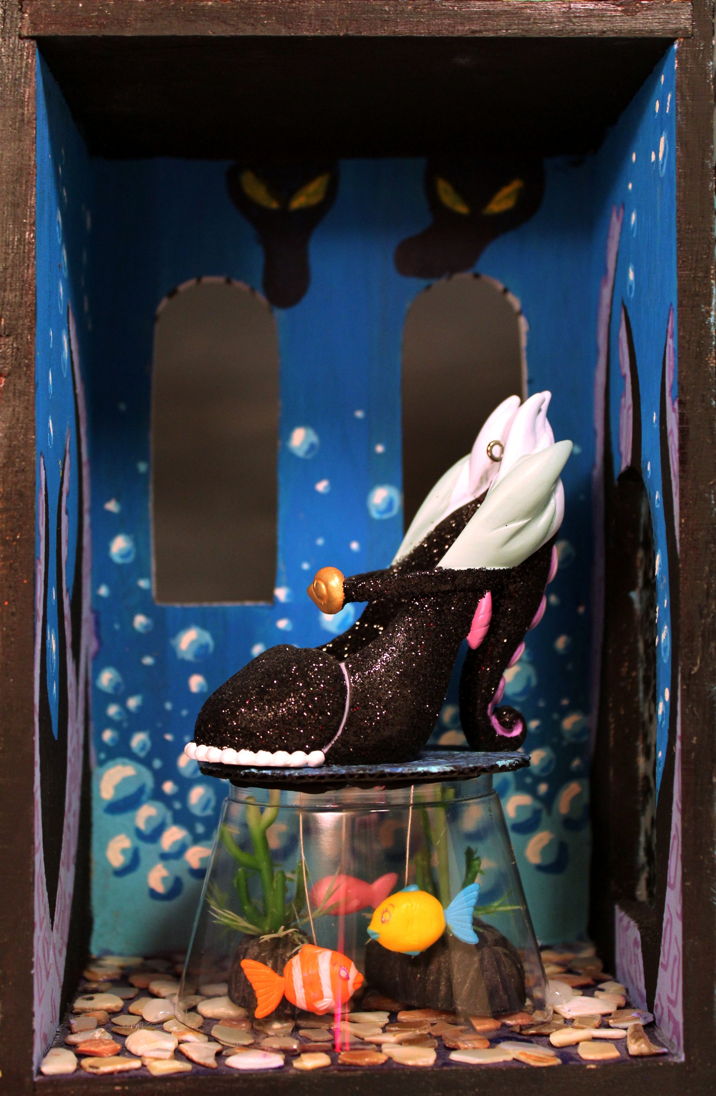 A black cat figurine with a pink tail, white paws, and a gold bell on its collar, sitting on a platform inside a decorated box with a blue background, painted black cats, and stars. Below the platform, glass with artificial fish and plant decorations