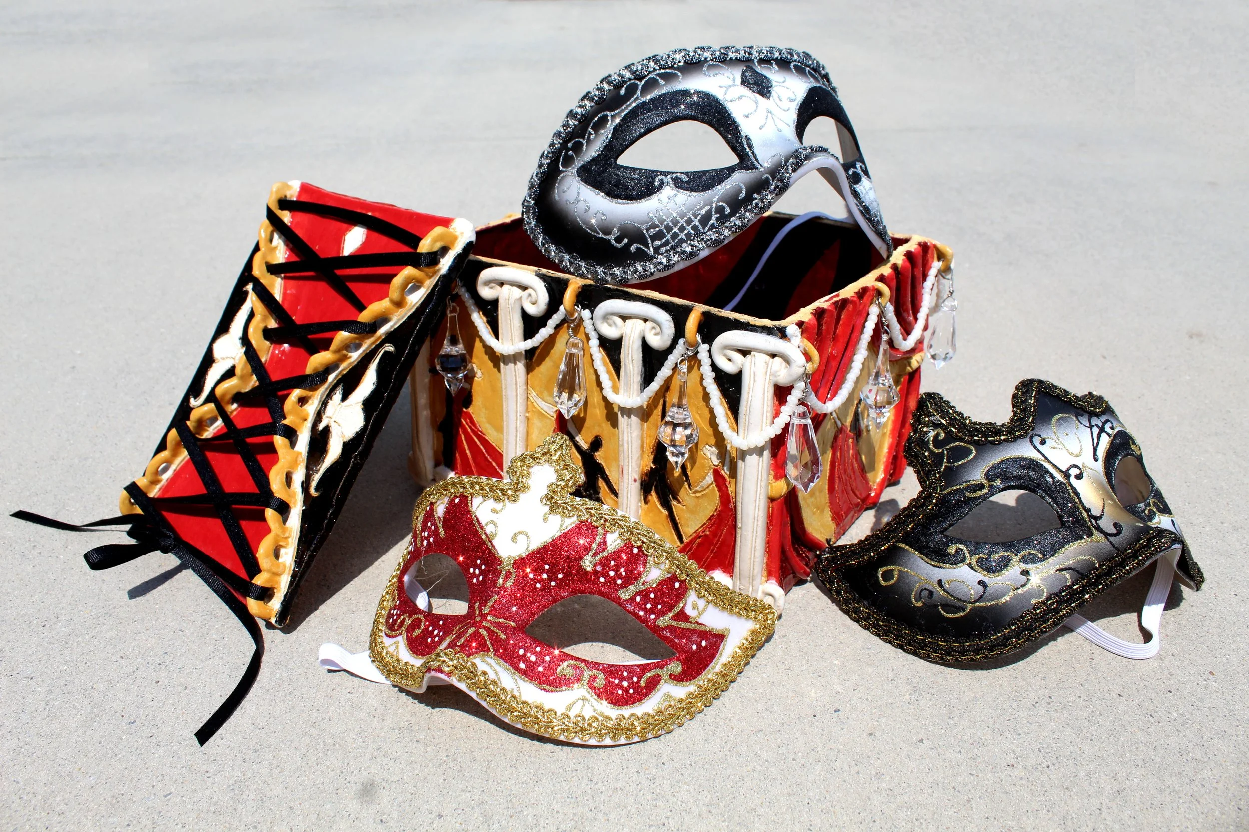 Multiple decorative masks and a miniature masquerade mask are arranged on a light-colored surface. The masks are designed for festive or costume use, featuring various colors and ornate details, including glitter, beads, and patterns.