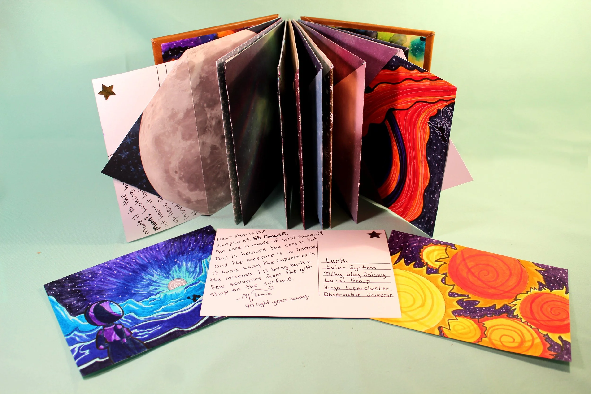 A collection of colorful illustrated cards featuring space, planets, and cosmic themes, arranged in a fan shape, with handwritten notes on some cards, displayed on a light blue surface.