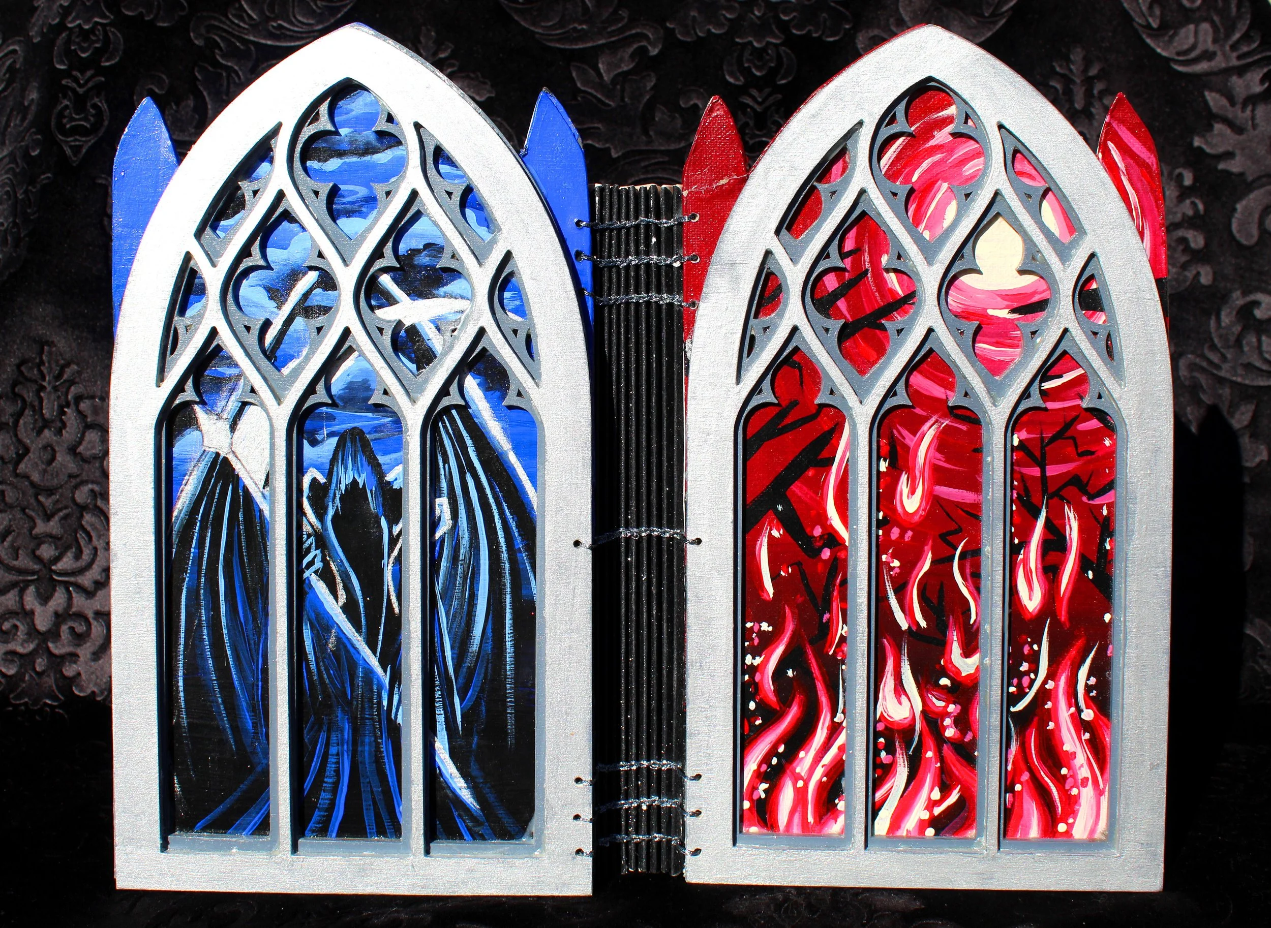 A decorative open book-shaped card featuring stained glass-style artwork with blue and red color schemes. The book is placed on a dark, patterned surface.