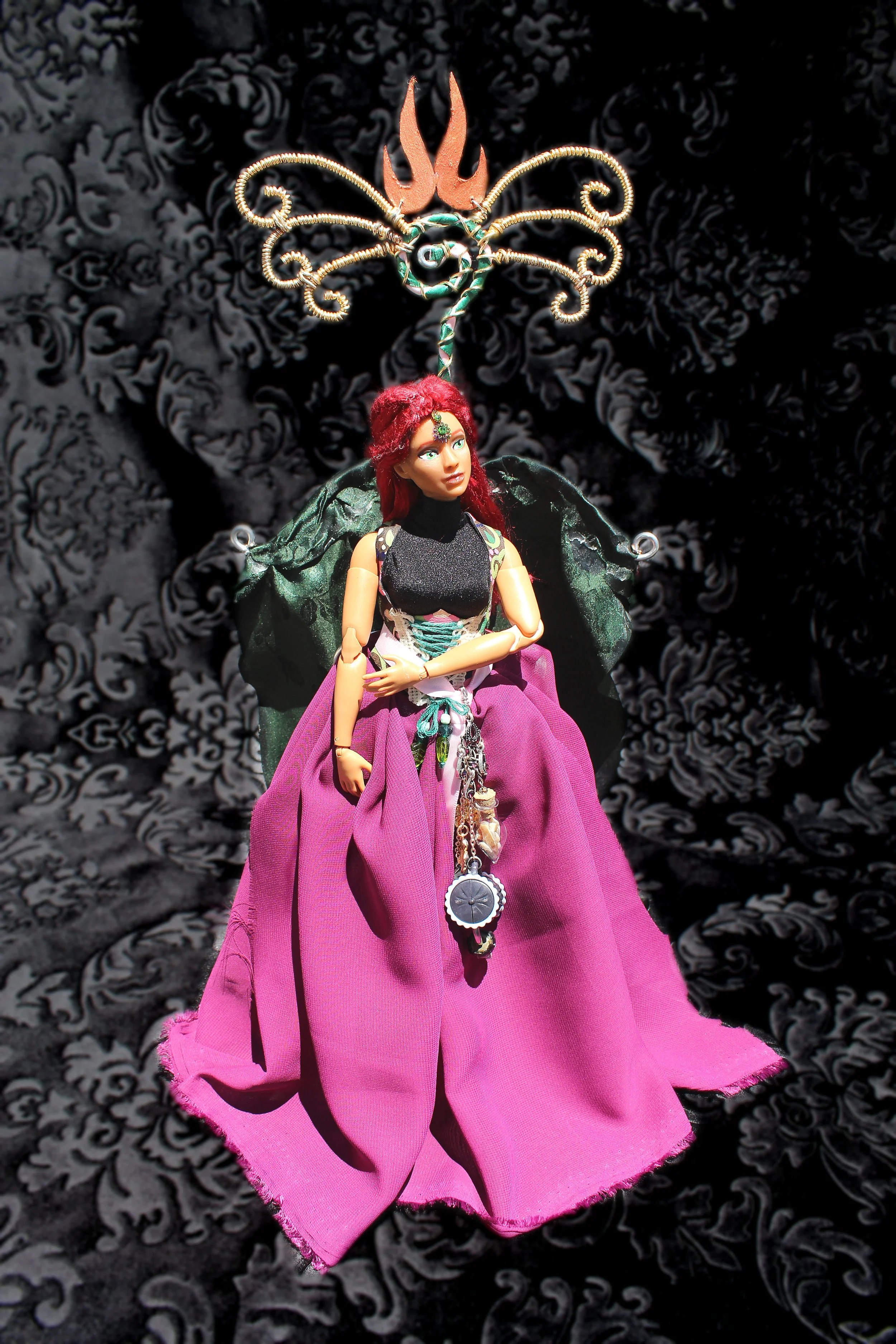 A doll with red hair, wearing a black sleeveless top and a long pink skirt, is showcased with butterfly wings and a decorative headpiece. The background is black with white intricate patterns.