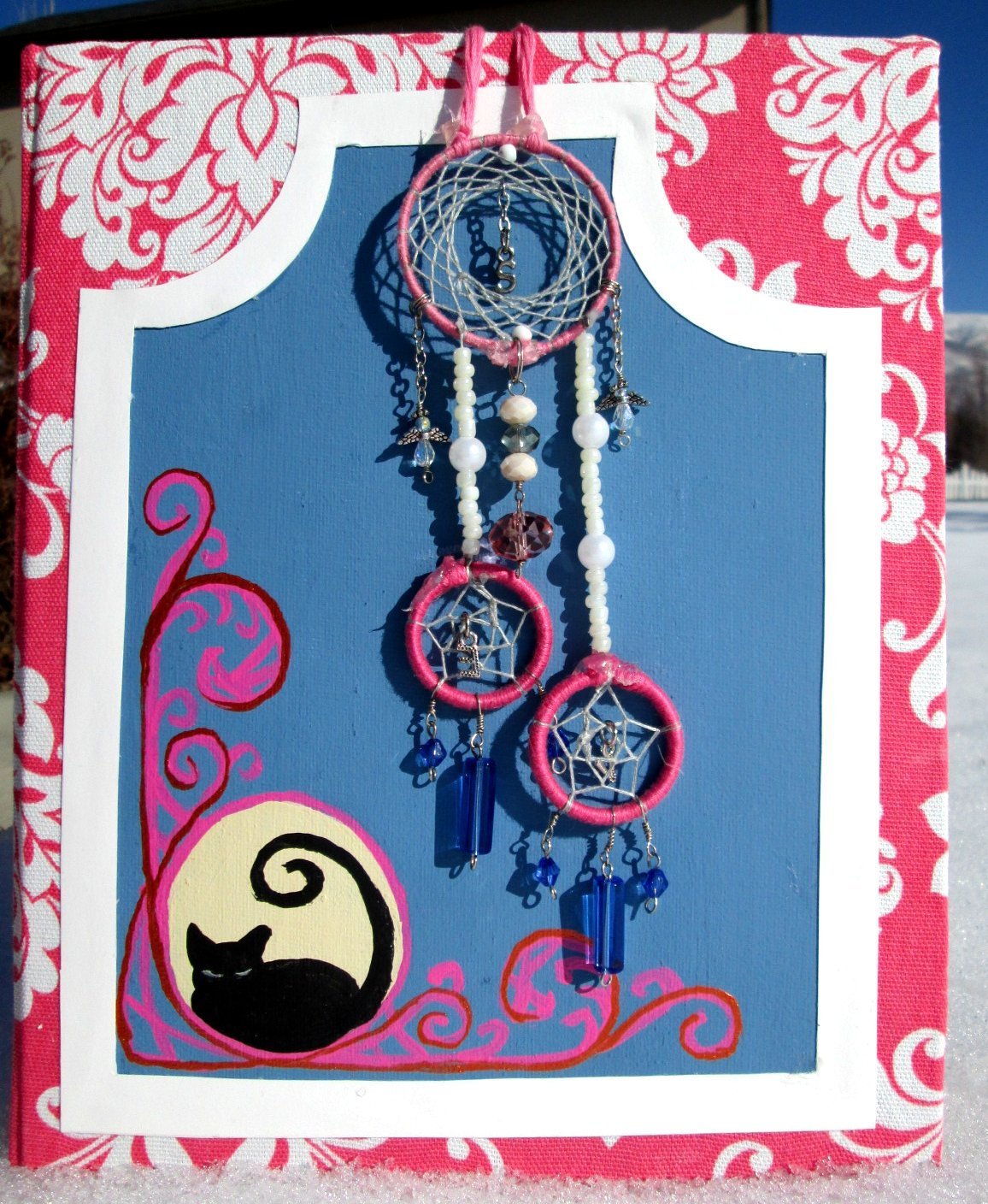 A decorative display of pink and white dreamcatchers with blue and clear beads, hanging against a blue background with a pink and white floral border and a black cat illustration.