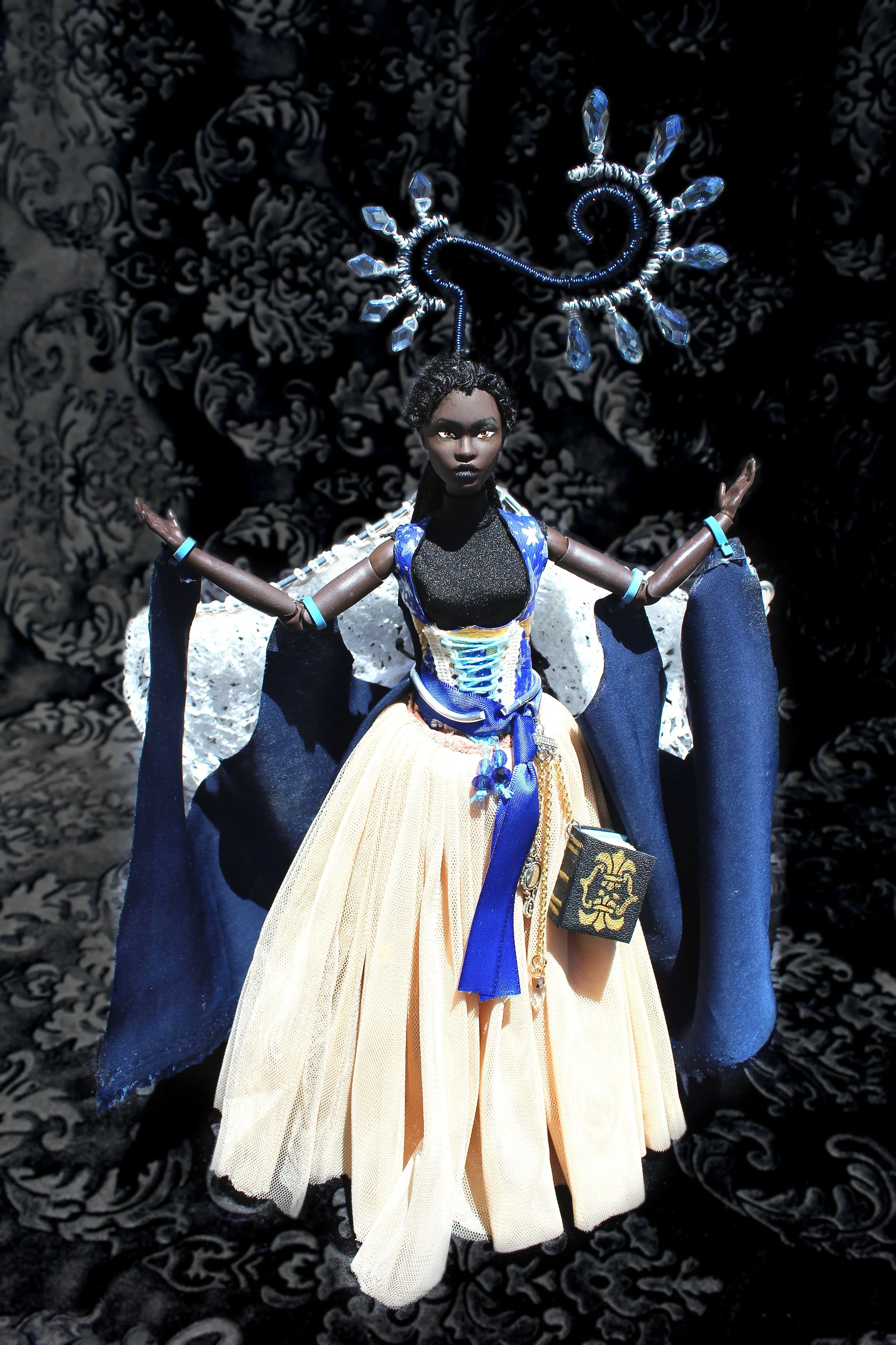 A doll dressed in elaborate costume with black skin, curly hair, and dramatic makeup. The doll has a large blue and gold headdress and wears a blue and gold gown with a yellow pleated skirt. The background holds a dark, patterned fabric.