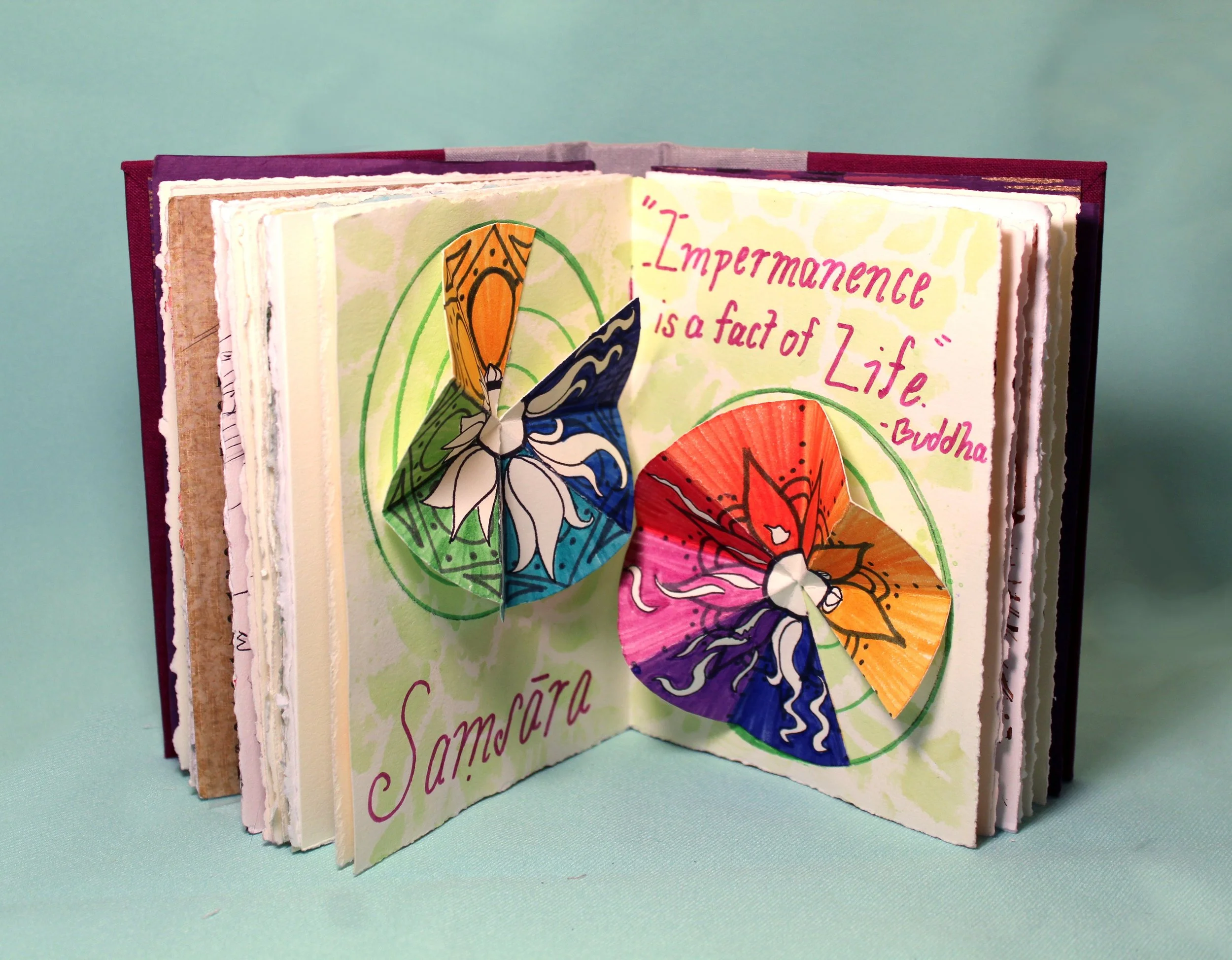 Open handmade paper booklet or art journal with colorful butterfly illustrations and pink handwritten text on a pastel green background.