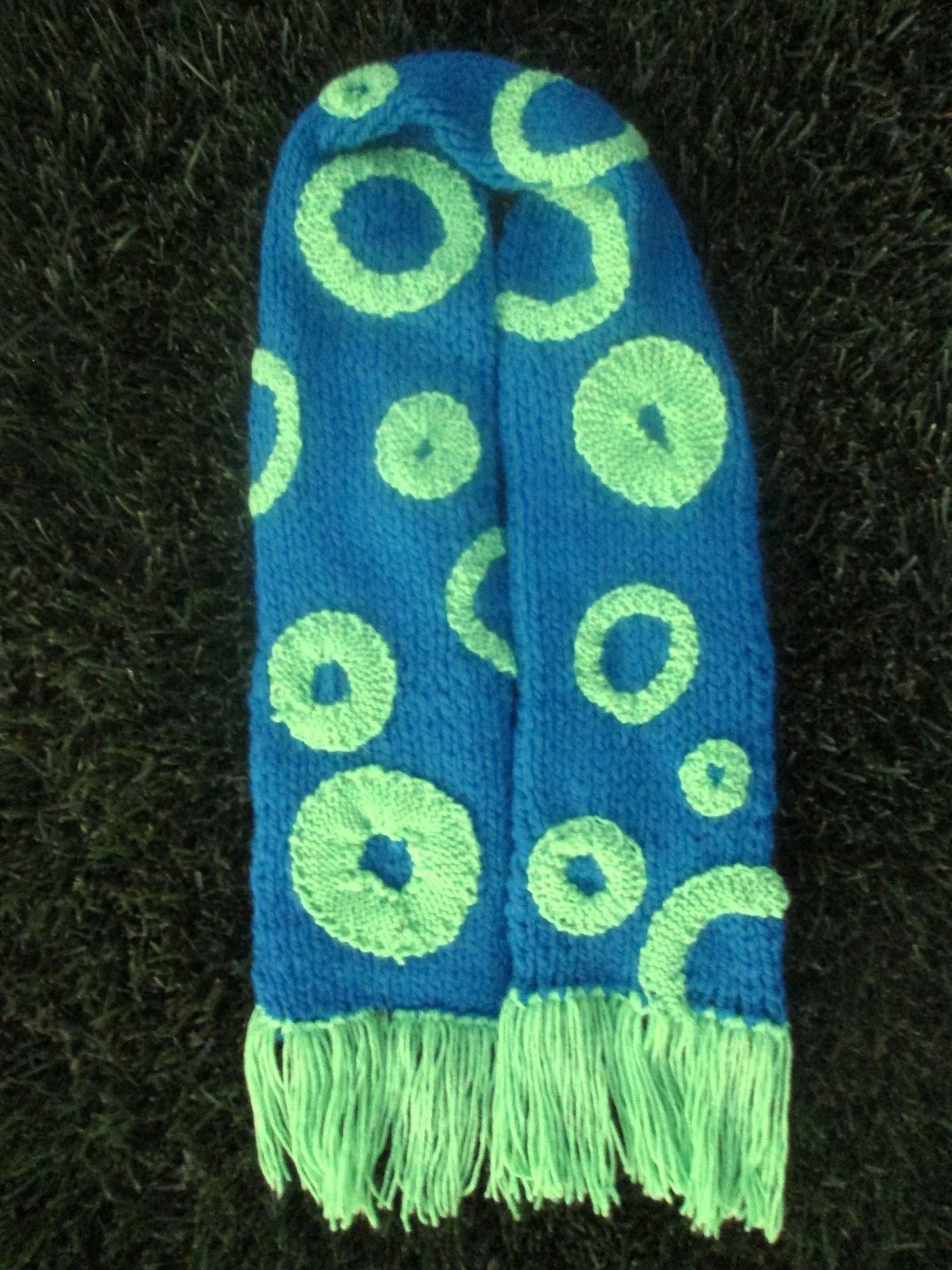 Blue knitted sock with yellow-green circle patterns and fringes on the toe and ankle, placed on grass.