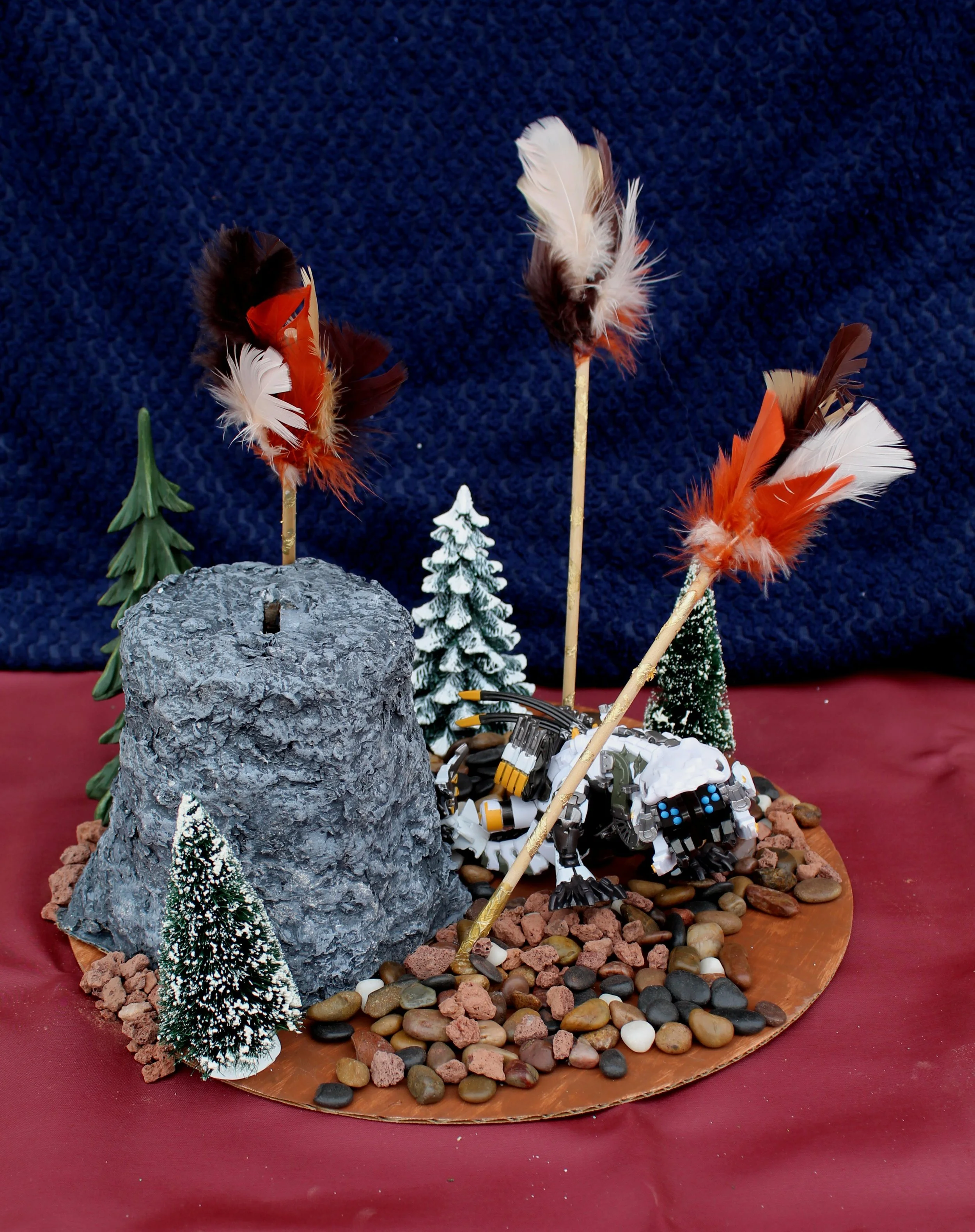 A diorama featuring a robot dog with feathers in a mountain scene with small trees, rocks, and pebbles on a circular base.