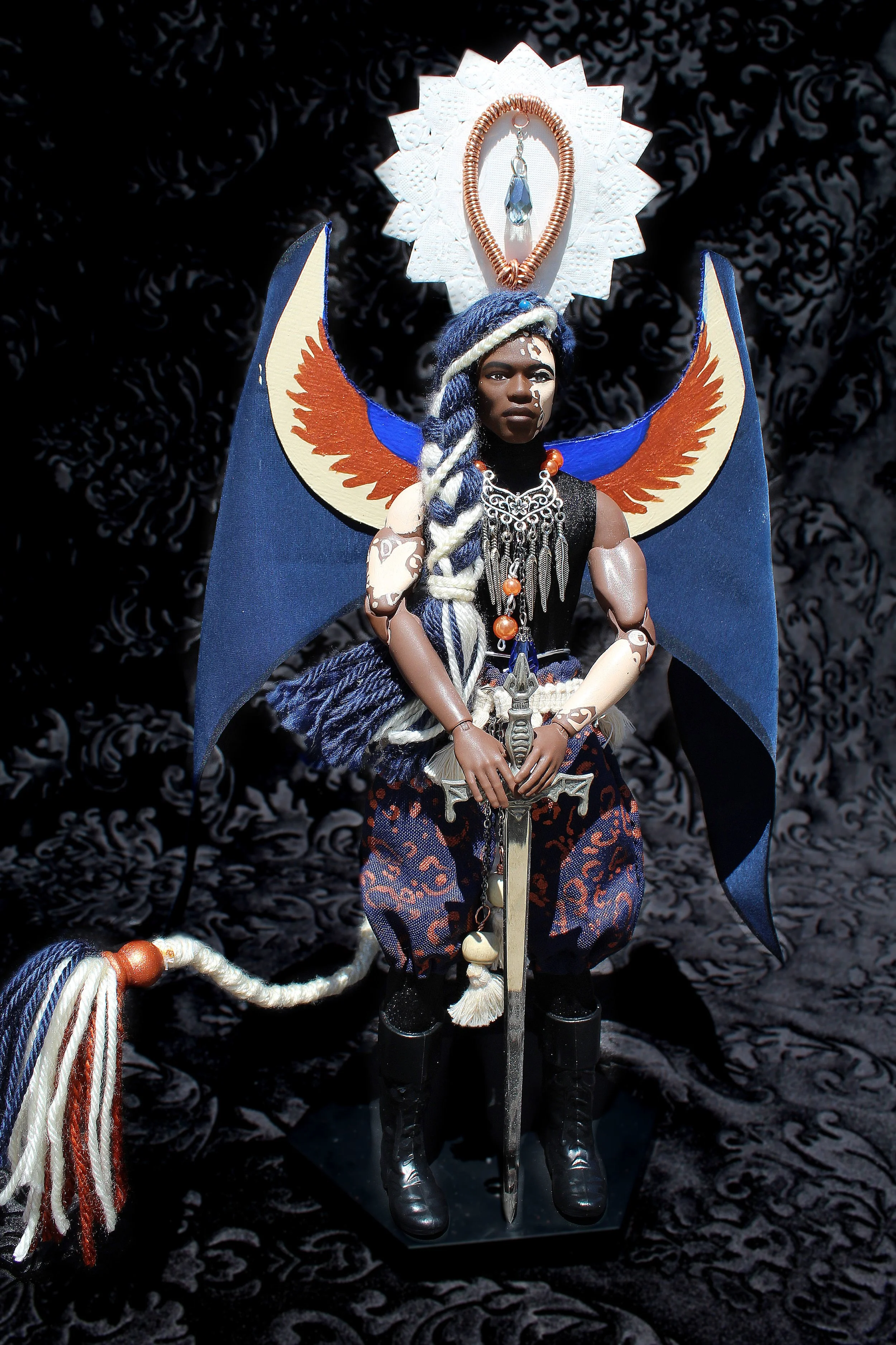 A doll dressed as a warrior or mythological figure with tribal and fantasy elements, holding a sword and adorned with jewelry and patterned clothing, standing against a dark patterned background.