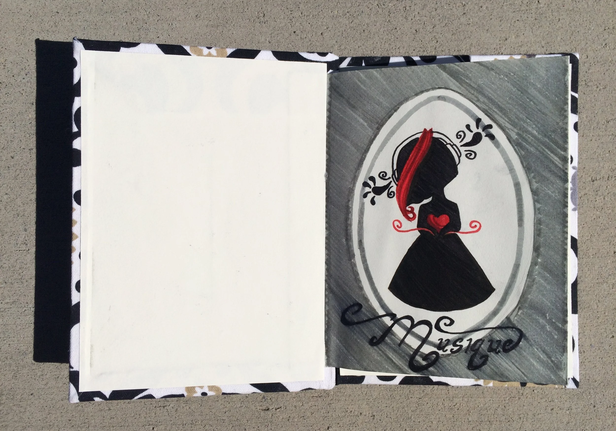 Open scrapbook with hand-painted design featuring a black silhouette of lips with a red ribbon, decorative swirls, and text that appears to say 'an en Vogue' or similar, on a beige surface.