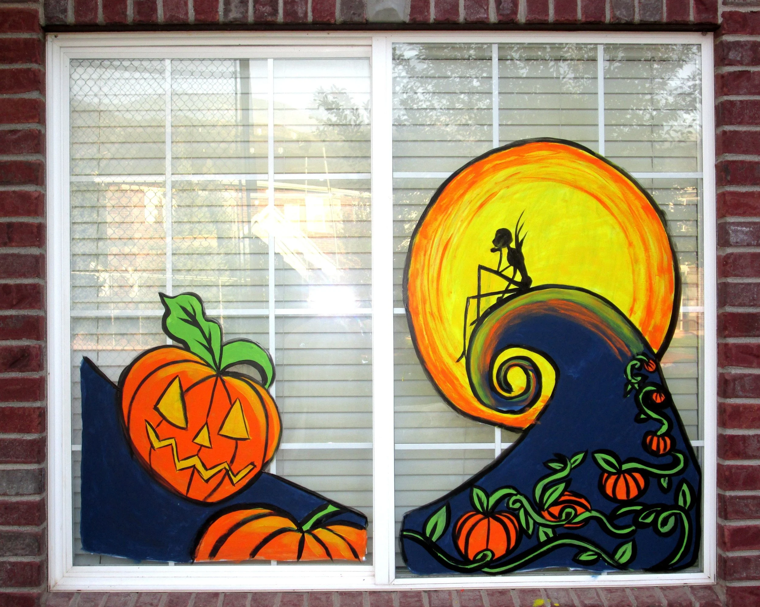 Colorful Halloween-themed window art featuring a smiling carved pumpkin, a black silhouette of a witch reading a book inside an orange moon, surrounded by green leaves and small pumpkins.