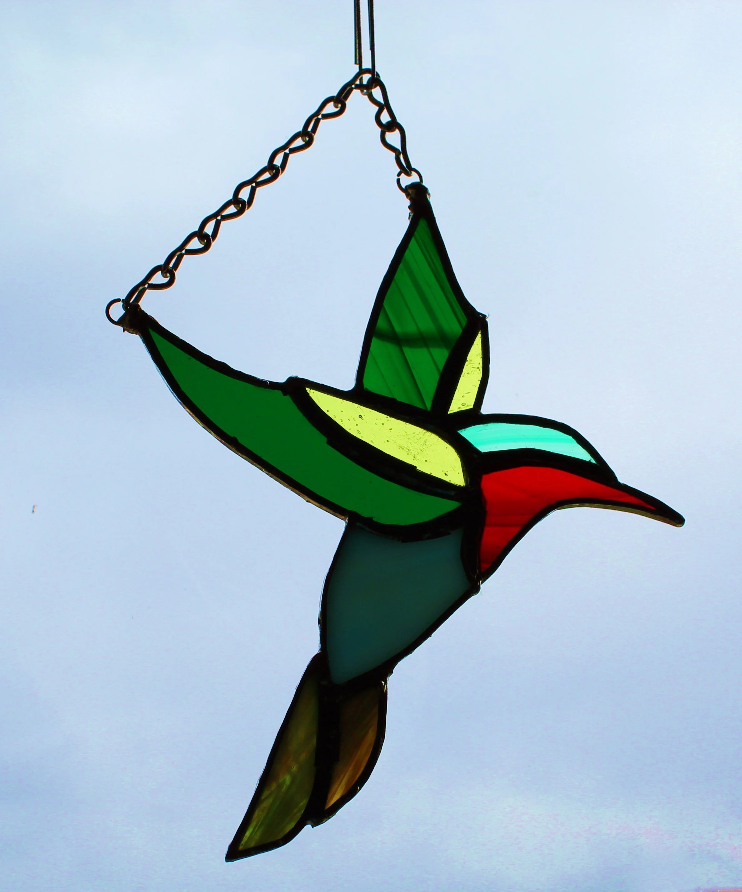 A stained glass sculpture of a bird in flight with colorful glass panels and a metal chain hanging from it, set against a cloudy sky.