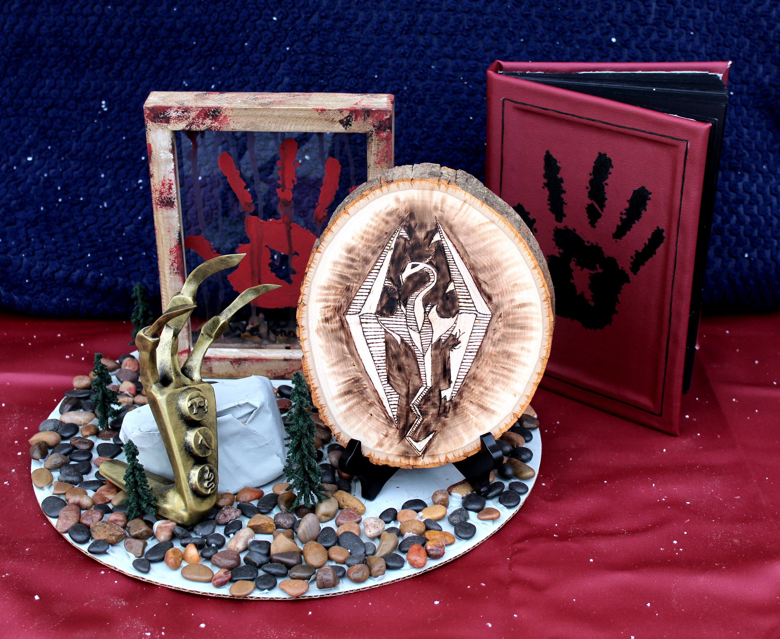 Artistic display featuring a painted wooden cube with red paint splatters, a wooden slice with black and white geometric painting, a bronze antler sculpture, a red framed artwork with black leaf design, and a backdrop of a dark blue textured curtain 