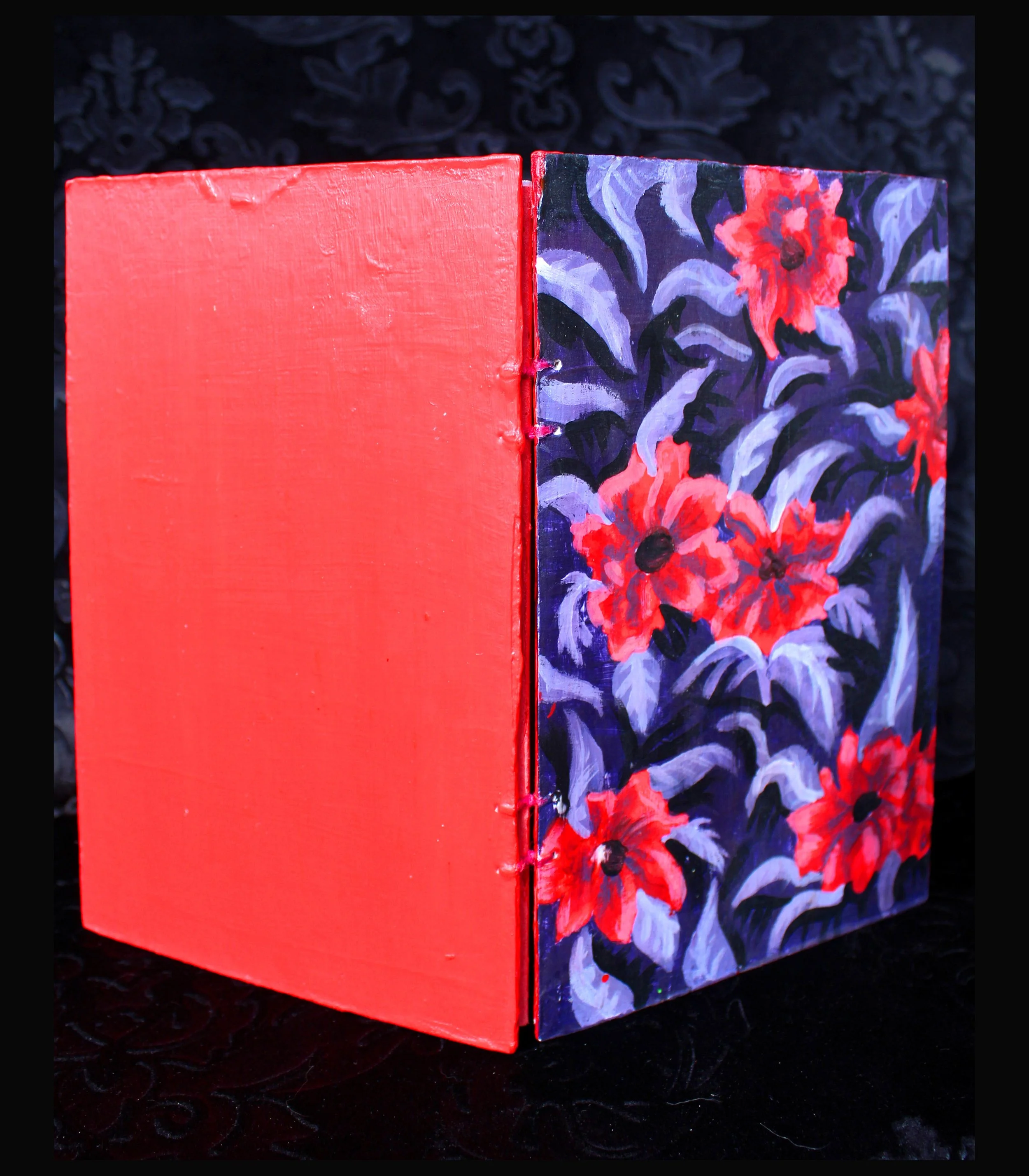 Close-up of a small book with a red spine and a cover painted with a dark background with red and lavender flowers.