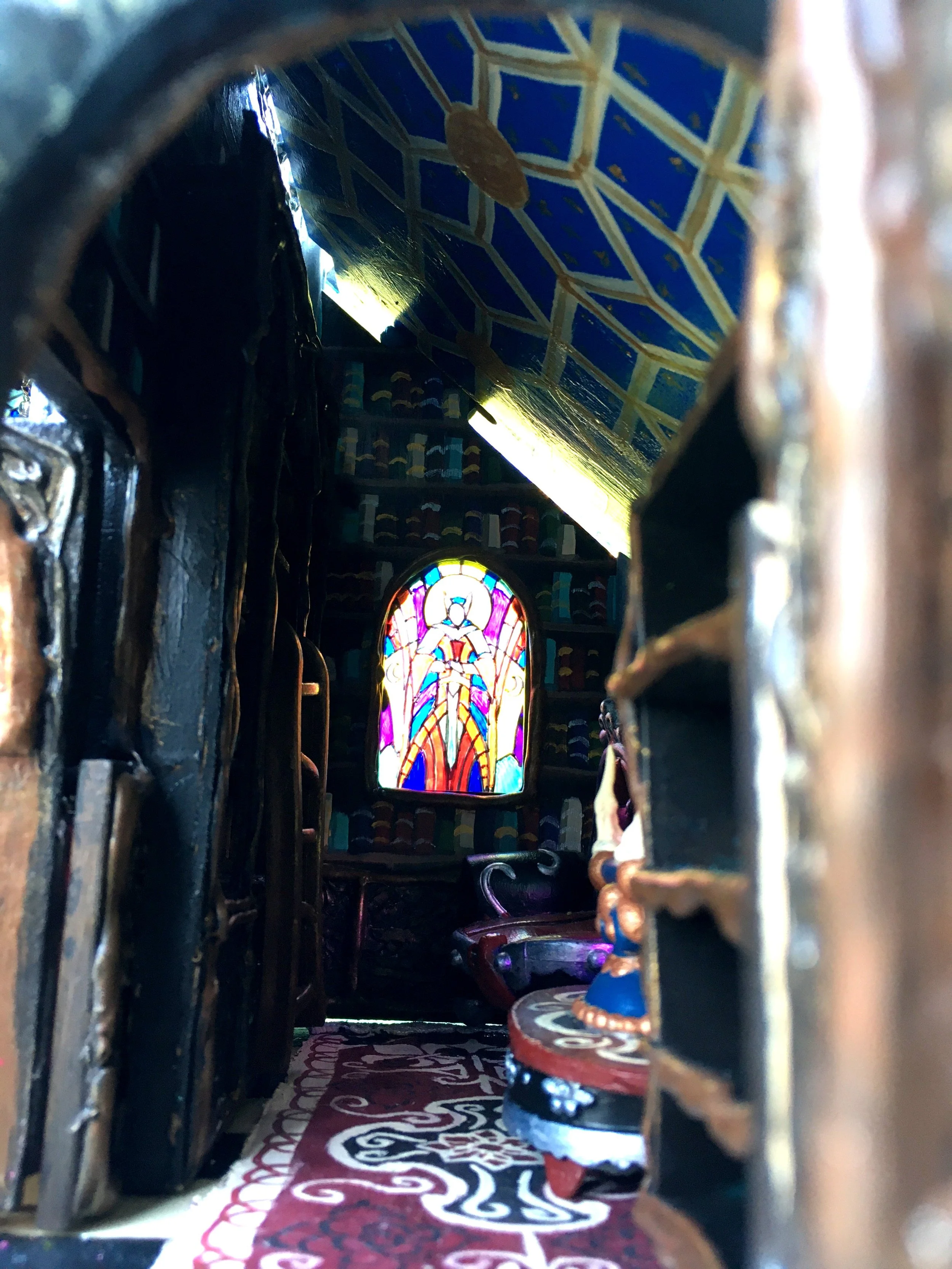 View of a colorful stained glass window with a biblical or religious figure, seen through a decorative iron or wood grille. The photo is taken from a low angle inside a religious or historical building, possibly a church or chapel.