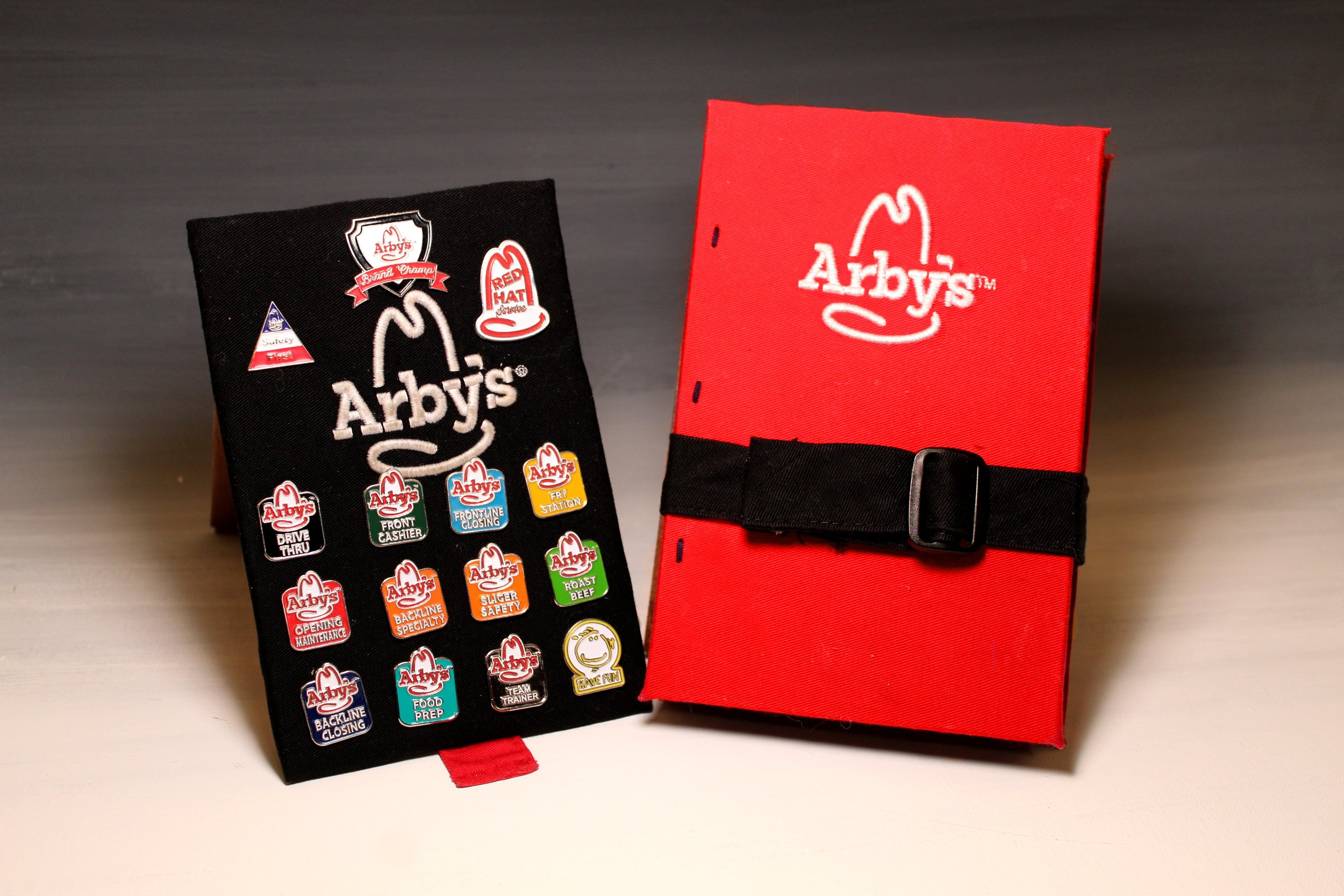A red Arby's hat stand with a black strap and a black Arby's sticker with various serviced-related icons and labels.
